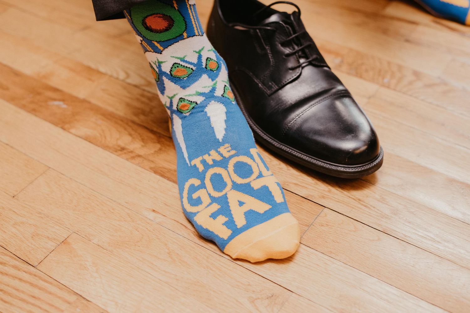 Close-up of a man wearing a blue 'The Good Fat' novelty sock with a black dress shoe on a wooden floor.