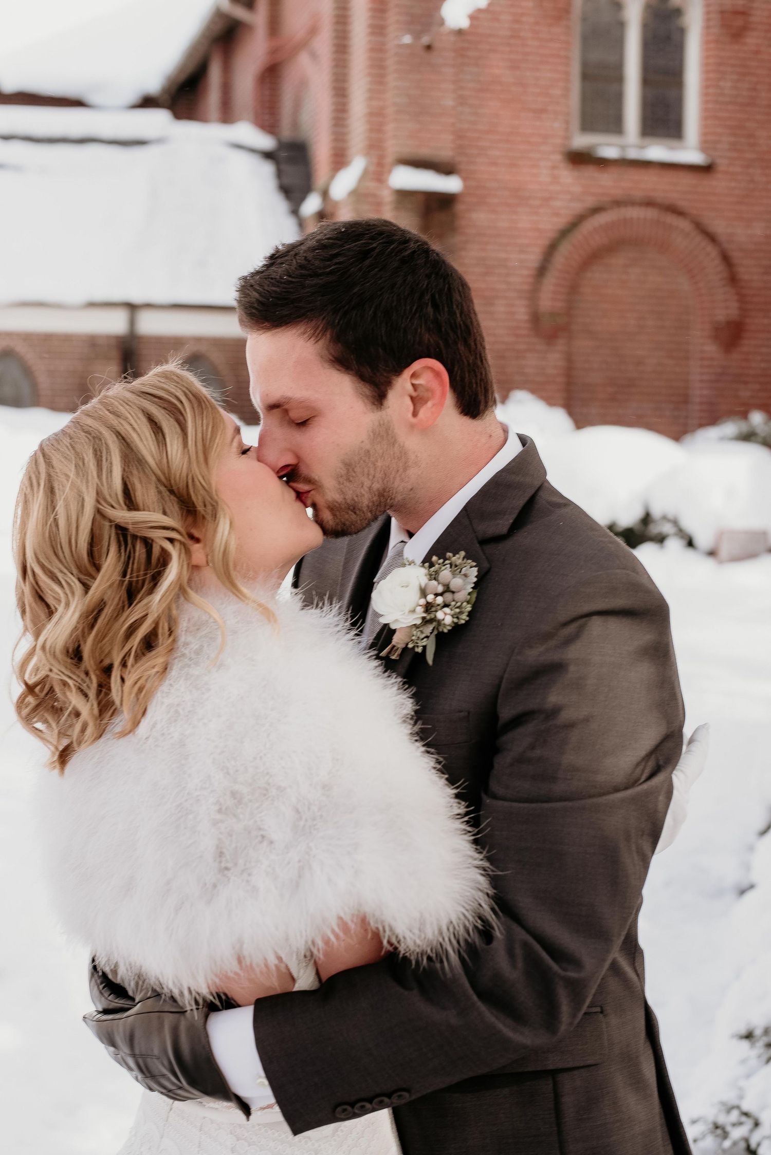 Bride and groom share a romantic winter kiss outdoors, bride wearing white fur wrap, groom in gray suit with boutonniere.