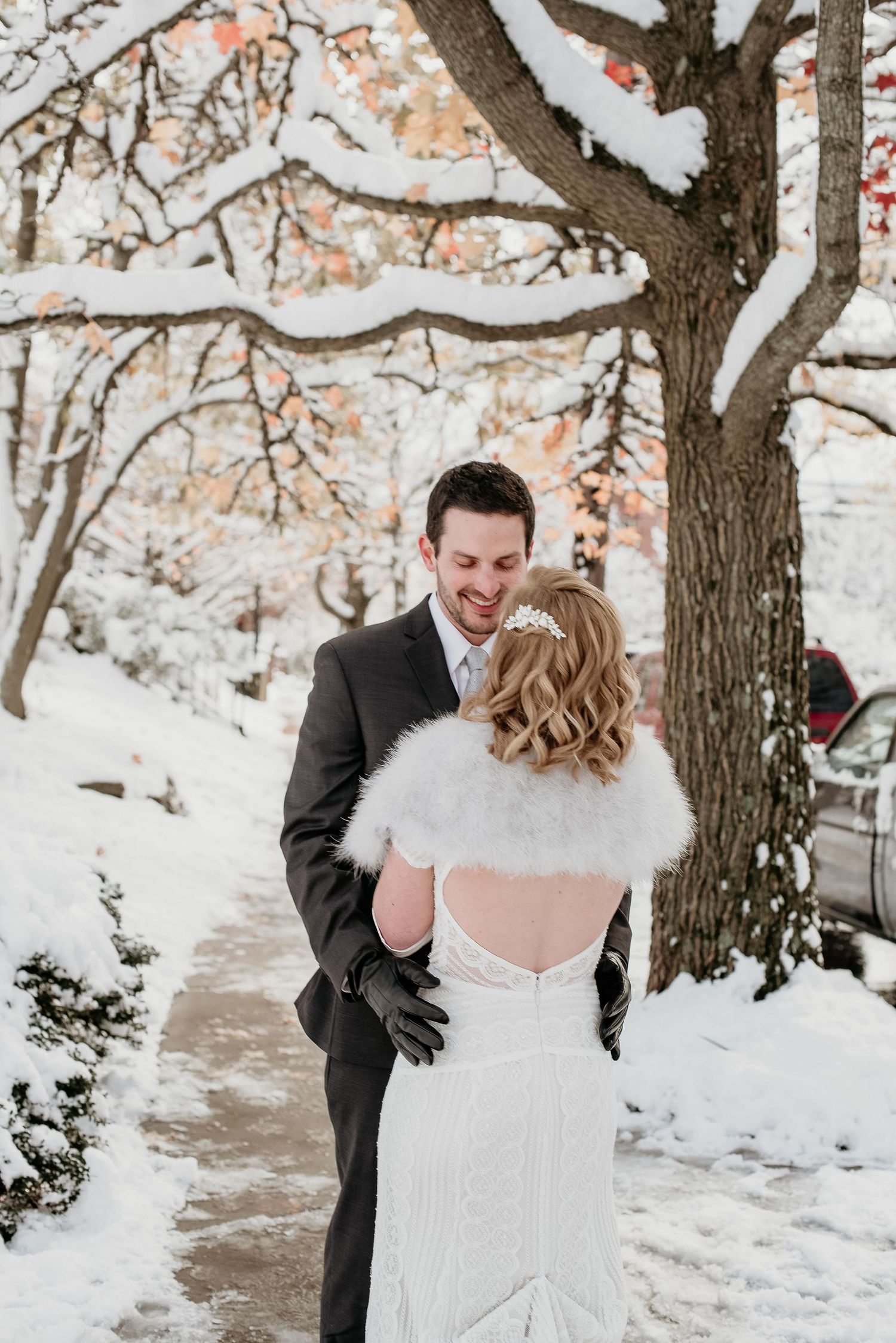 Smiling groom in dark suit with bride in backless lace dress and fur wrap during snowy winter wedding.