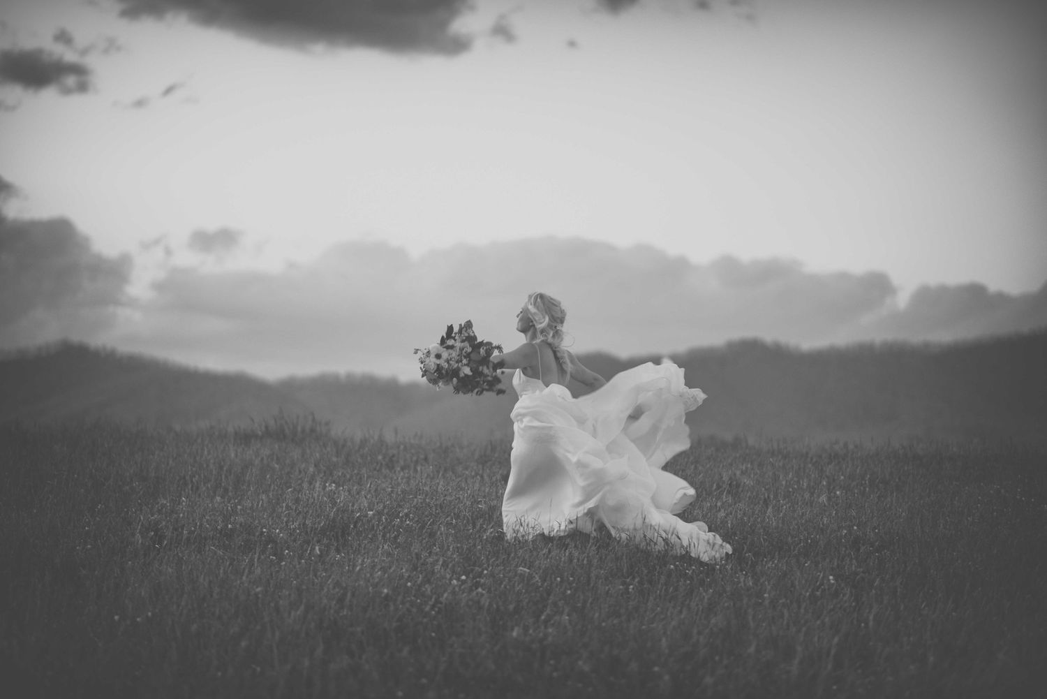 Black and white photo of a bride in a flowing white dress running through a grassy field holding a bouquet.