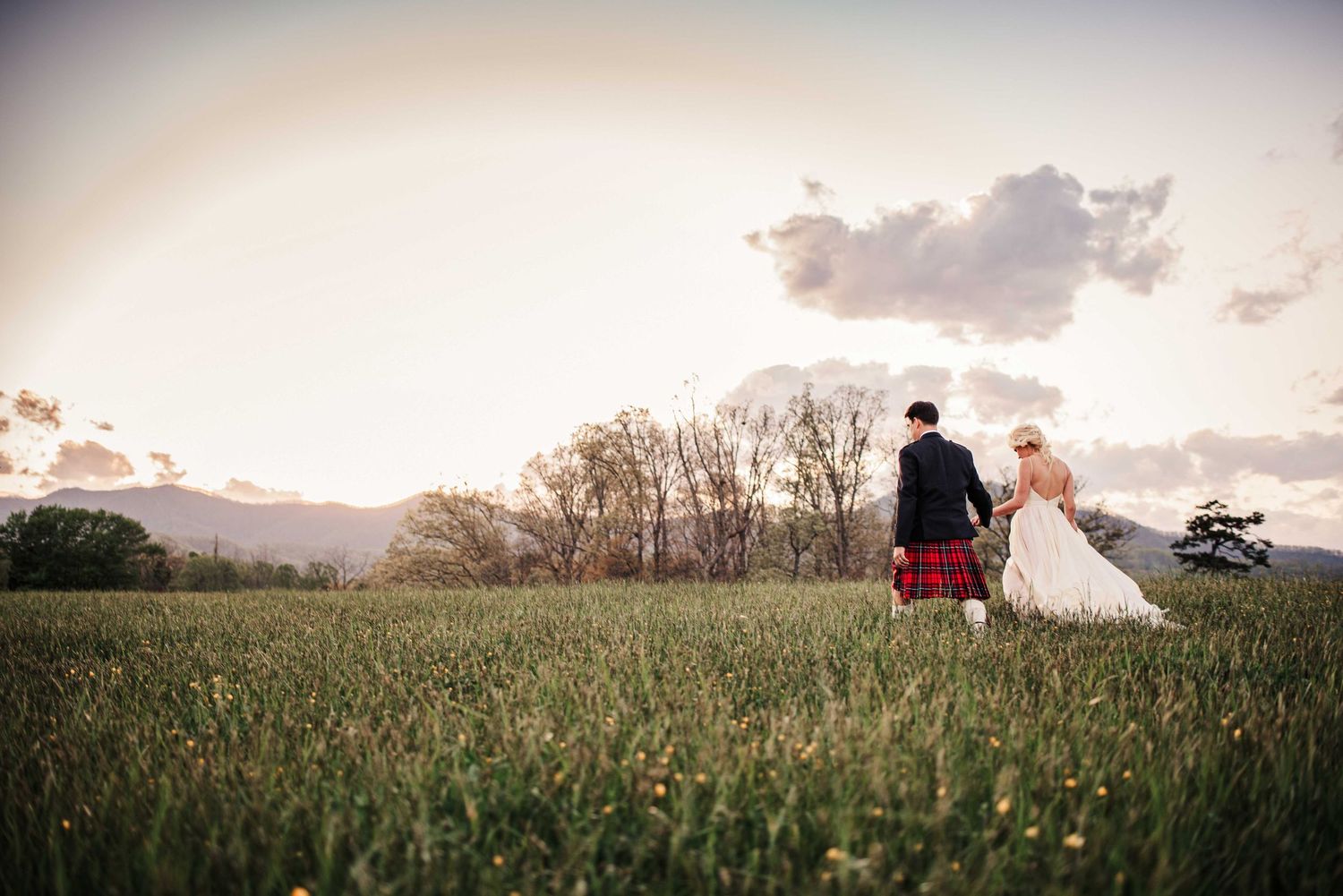 A bride and groom walk hand in hand through a lush green field at sunset with trees and mountains in the background.
