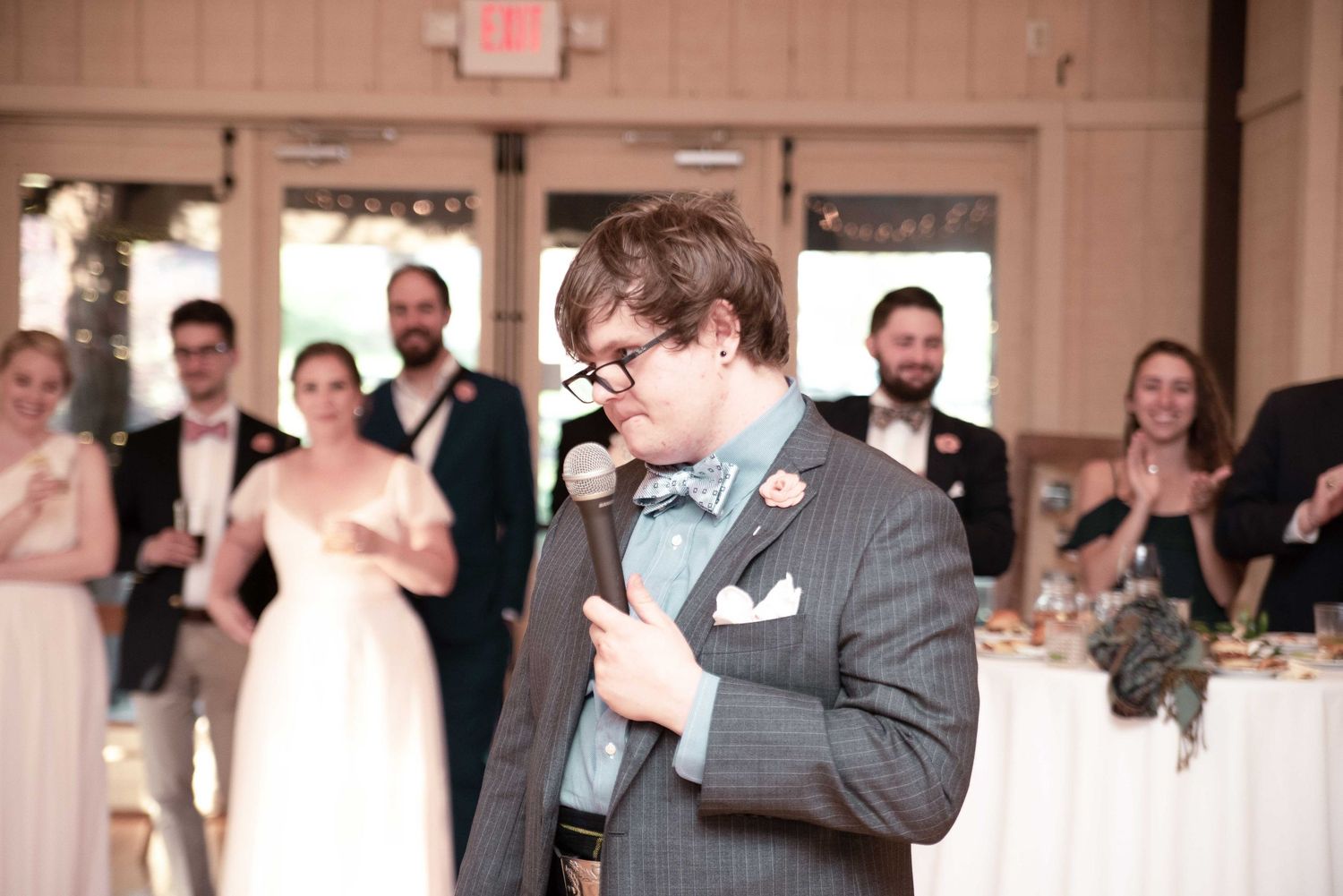 Young man in gray suit giving a speech at a wedding reception, holding a microphone with guests watching.