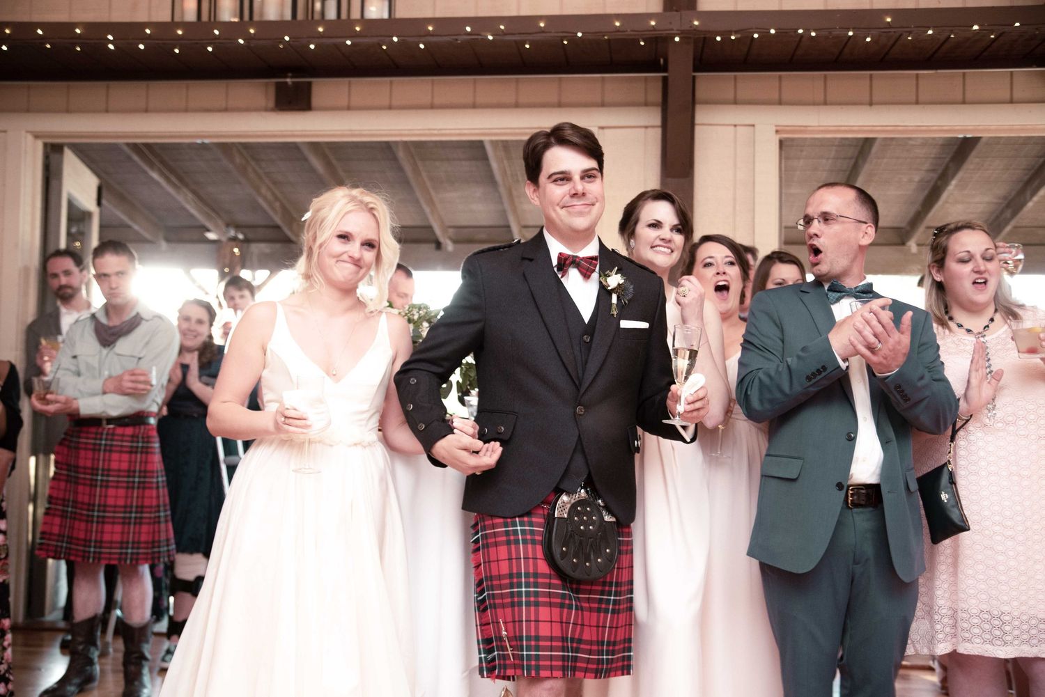 Bride and groom in Scottish kilt celebrate at wedding reception with smiling guests clapping around them.