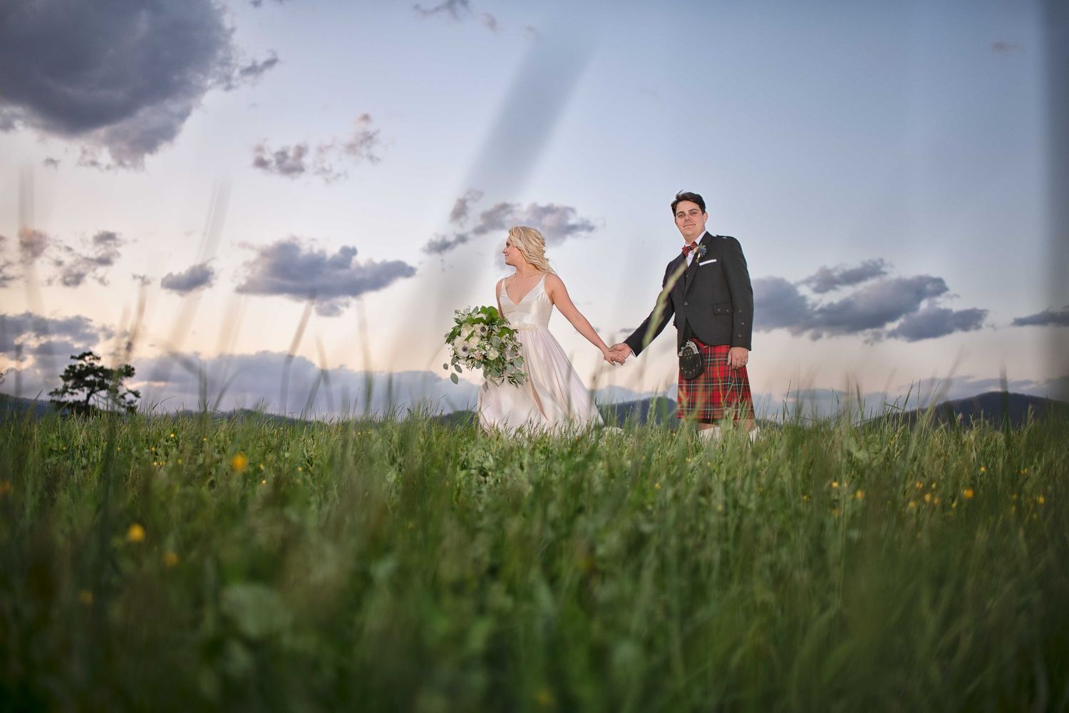 Bride and groom holding hands walking through green meadow at sunset with dramatic cloudy sky backdrop.