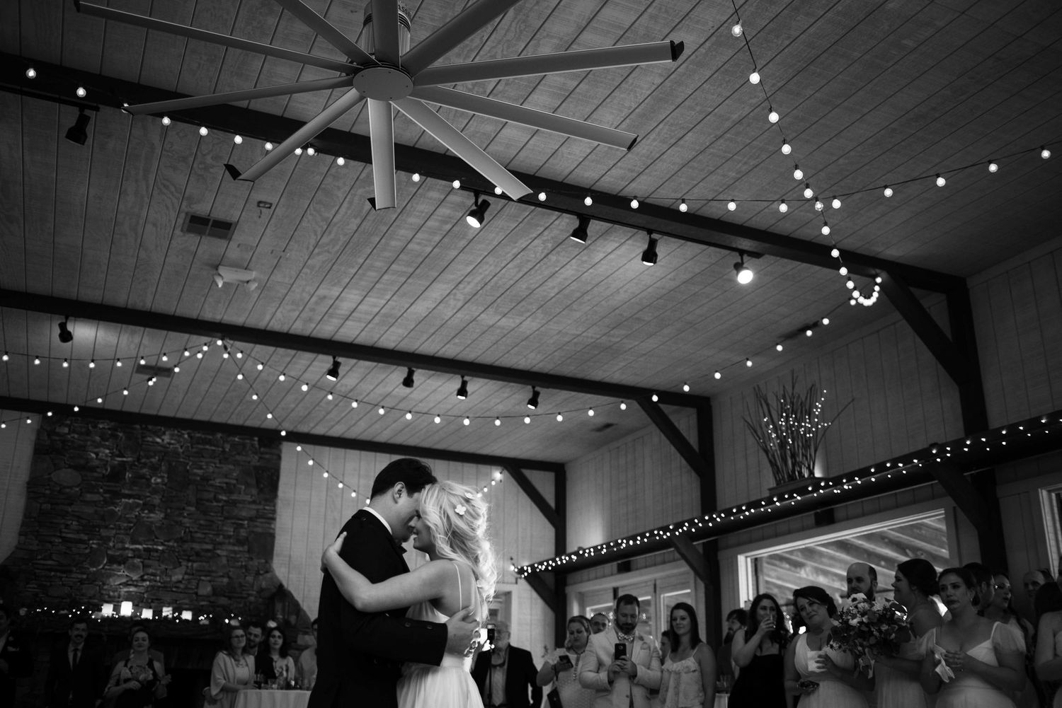Black and white photo of bride and groom sharing first dance under string lights in a rustic barn wedding venue.