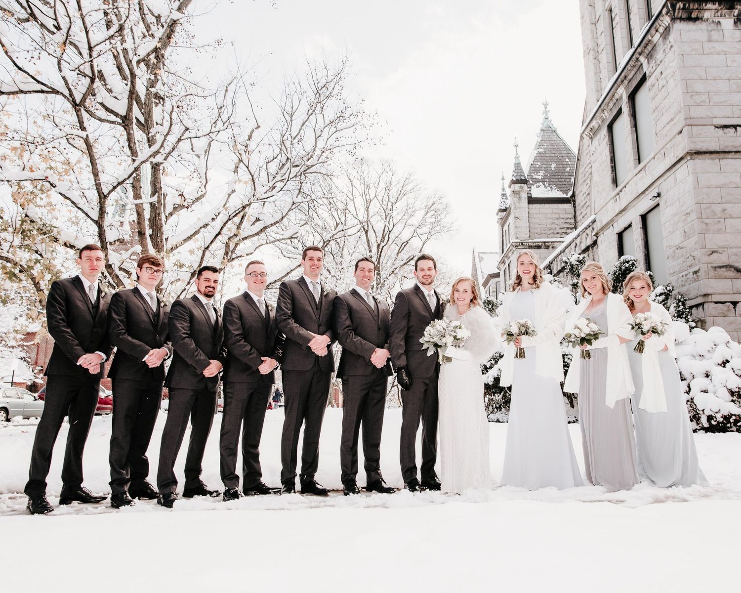 Winter wedding party standing outdoors in snow, men in dark suits beside bridesmaids in elegant gray dresses.