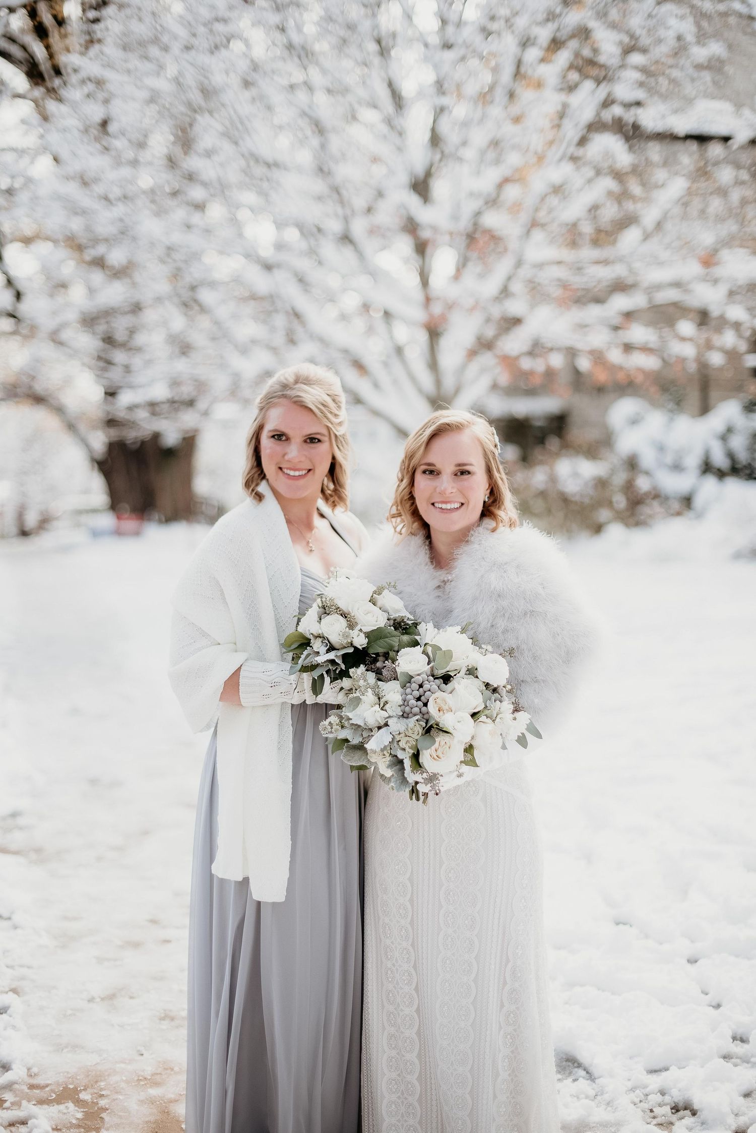 Two brides in white and gray gowns with fur wraps hold a white bouquet outdoors in a snowy winter wedding setting.