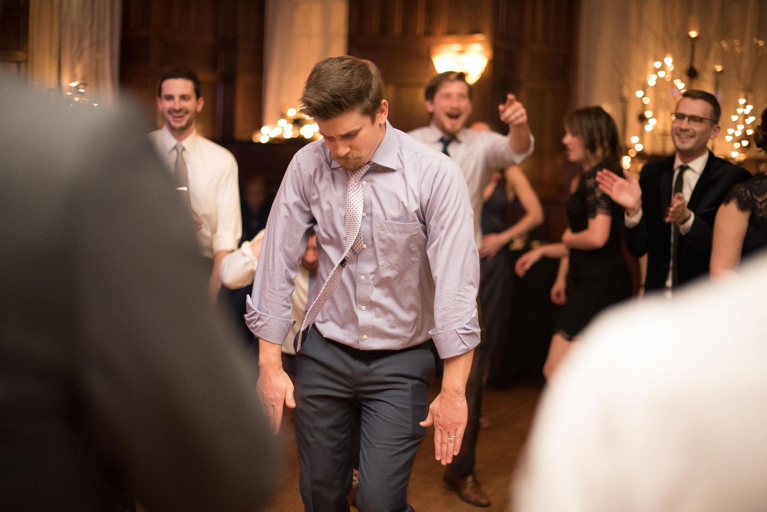 Man dancing at wedding reception holding microphone, guests laughing and celebrating in elegant ballroom background.
