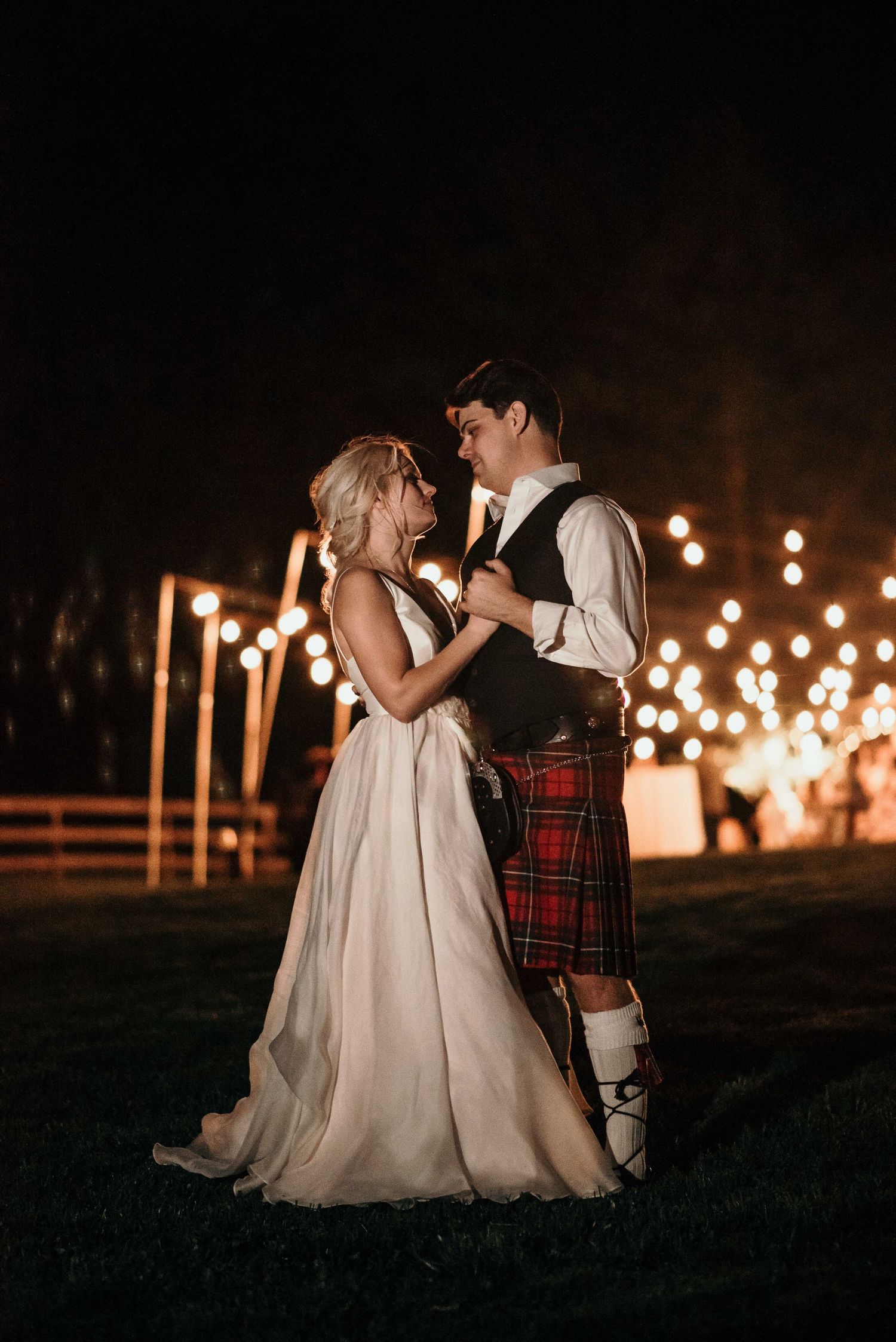 Bride and groom share first dance outdoors at night, groom wearing red tartan kilt, surrounded by warm glowing lights.