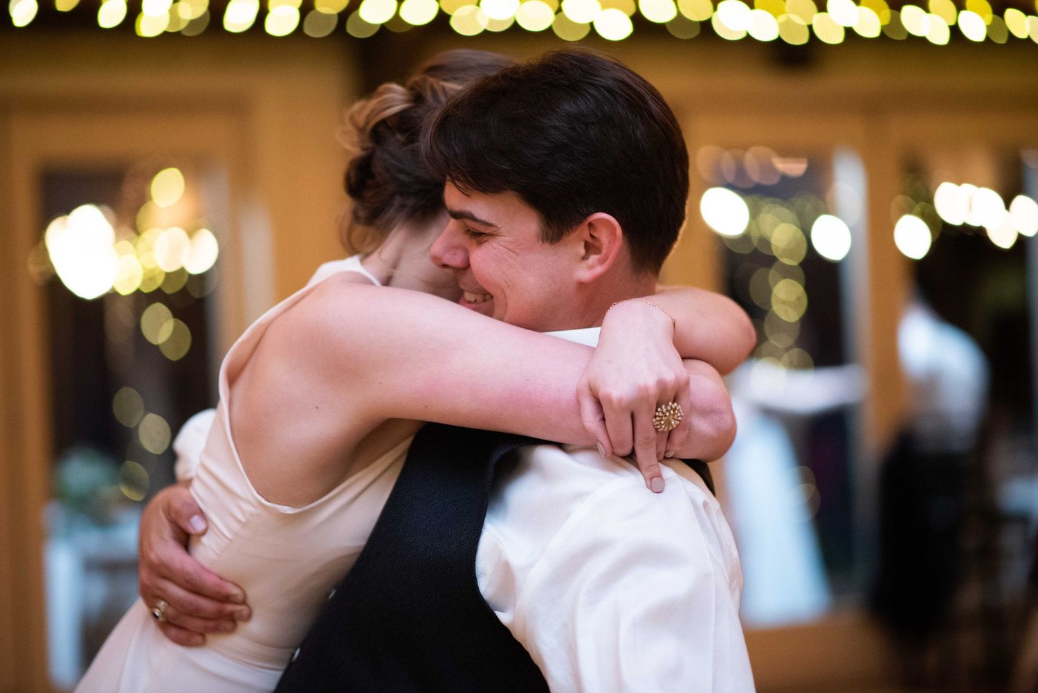A bride and groom share an emotional first dance hug at their wedding reception with warm bokeh lights in the background.