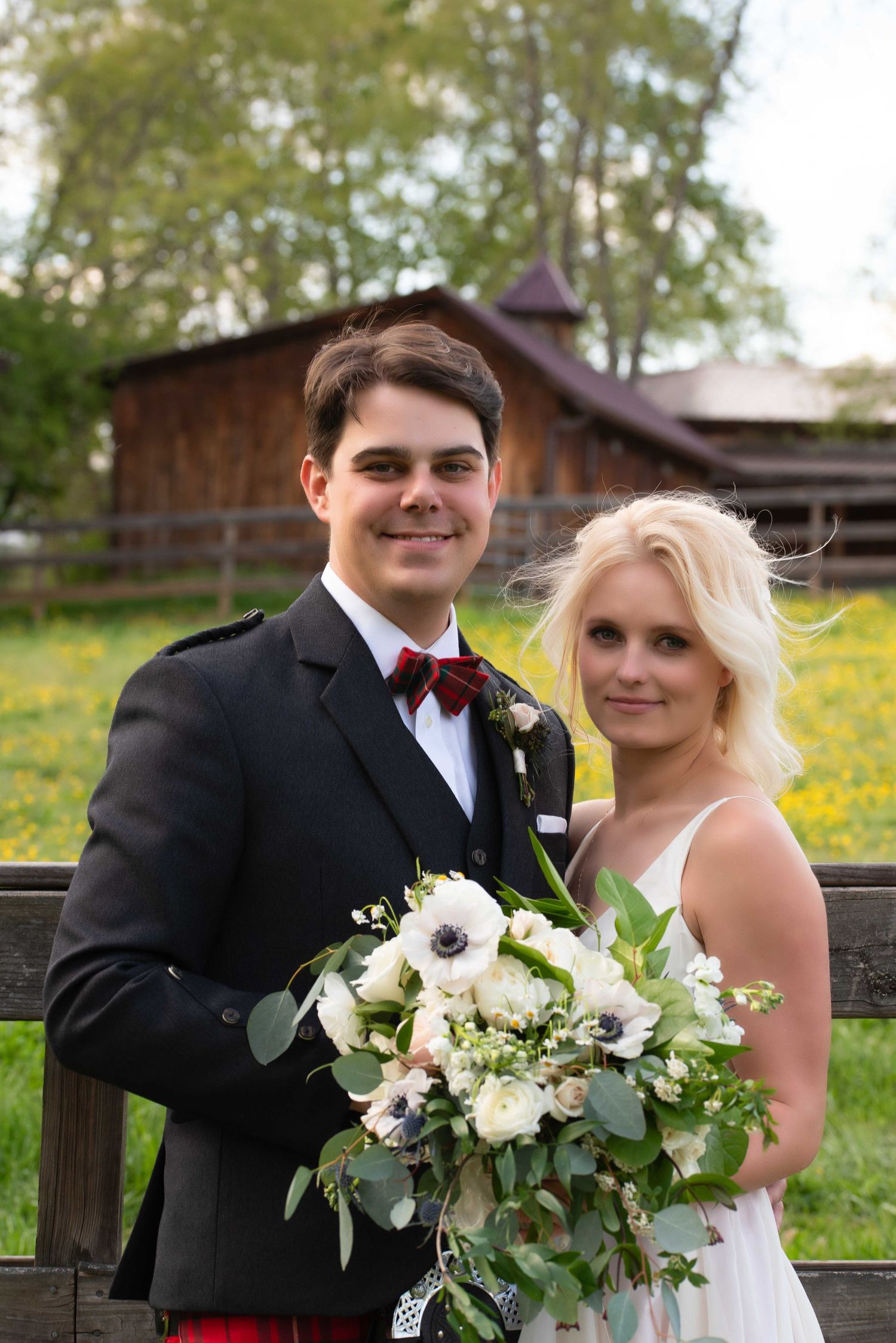 A smiling couple in wedding attire pose outdoors, groom in kilt with bow tie, bride holding white floral bouquet.