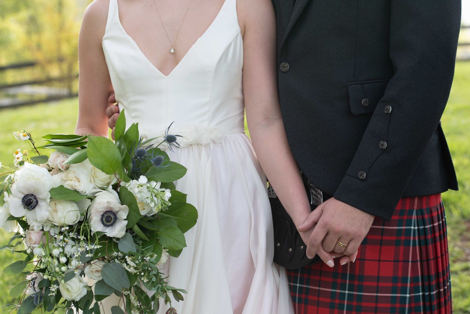 Bride in white gown holding white anemone bouquet stands beside groom wearing traditional red tartan kilt at outdoor wedding.
