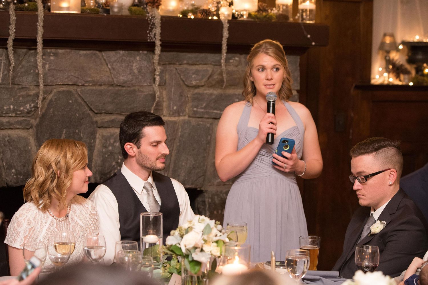 Bridesmaid giving a toast speech at a wedding reception near a stone fireplace.