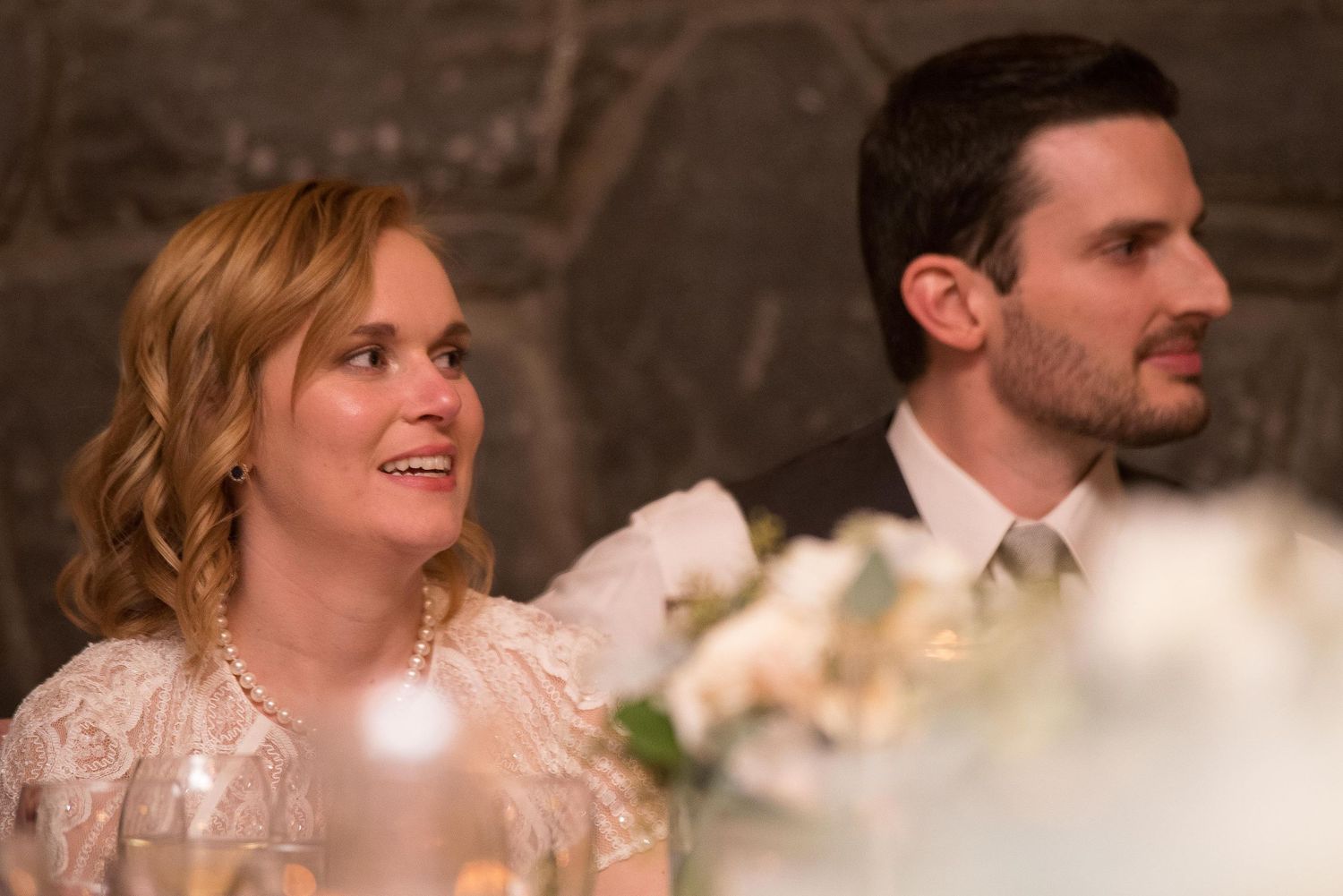 A woman in a white lace dress and pearl necklace sits beside a man in a suit at an elegant dinner event.