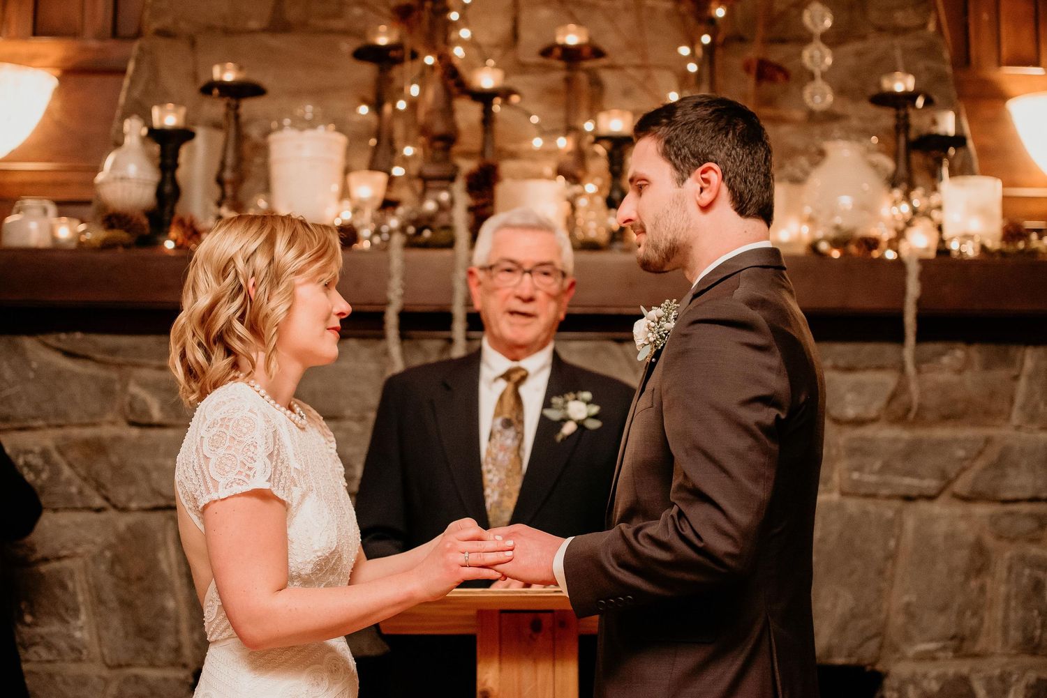 Bride and groom exchange vows at rustic stone fireplace wedding ceremony with officiant and candlelit backdrop.