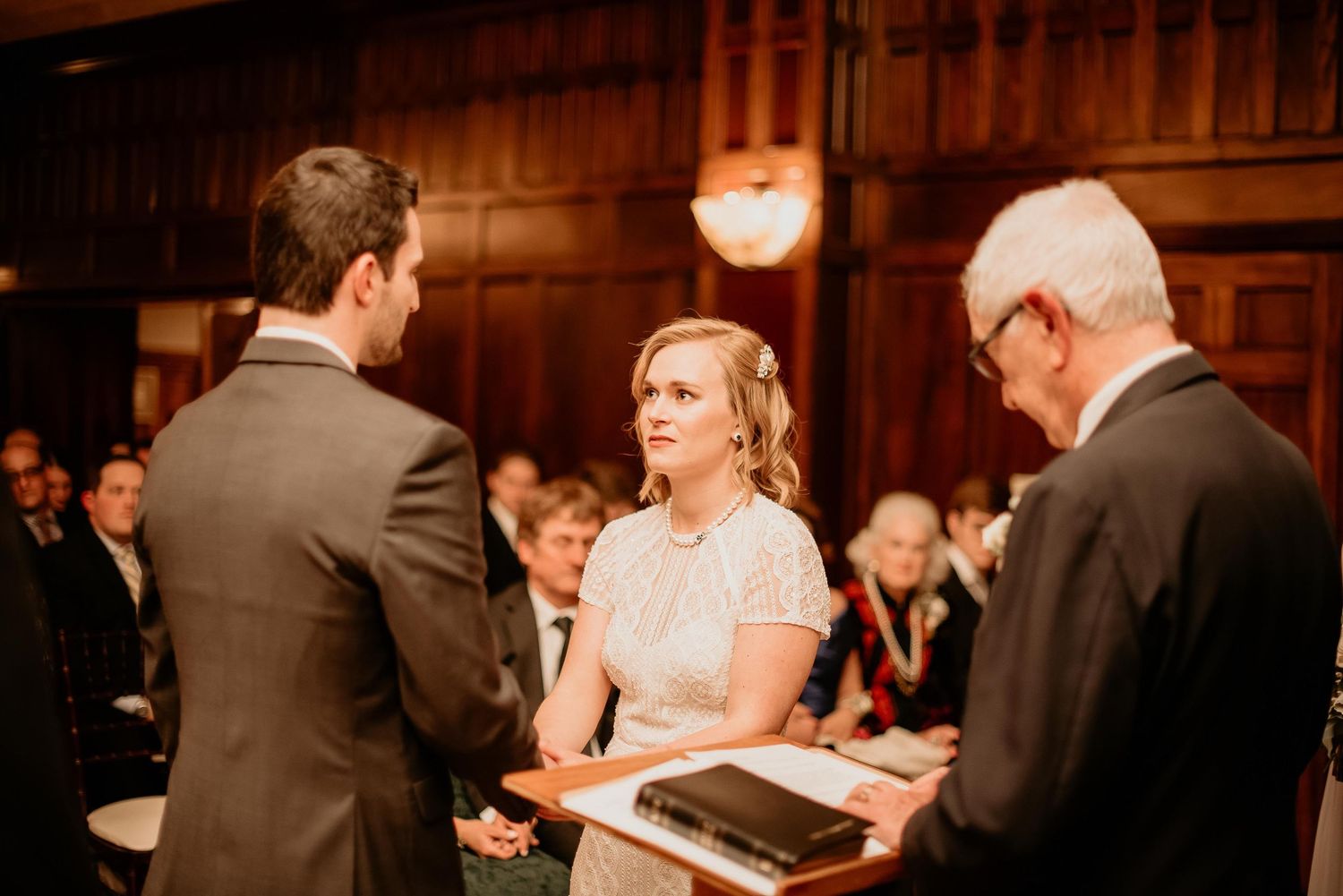 Bride and groom exchange vows at elegant wood-paneled wedding ceremony with officiant holding Bible.