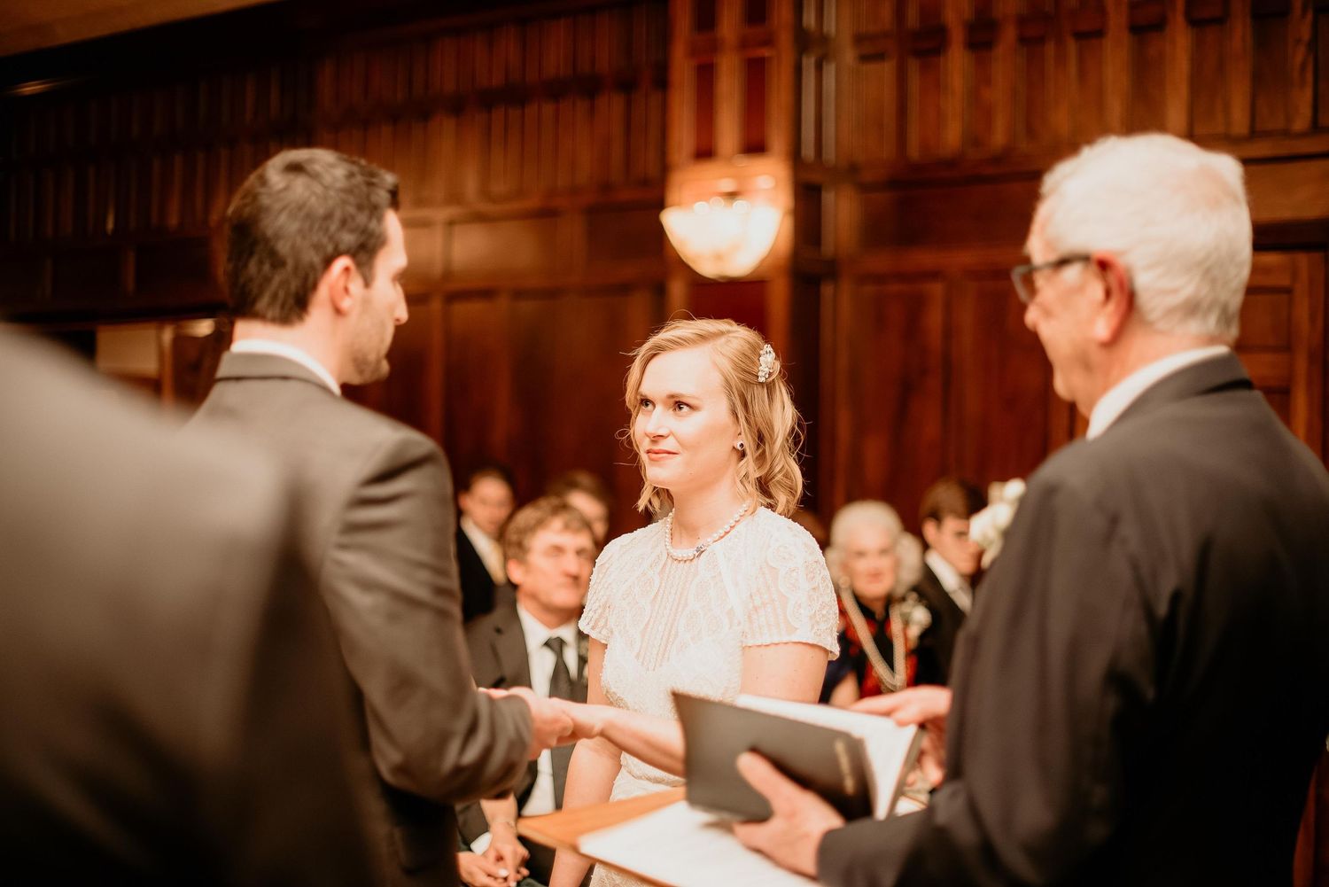 Bride smiling at groom during indoor wedding ceremony in elegant wood-paneled venue with guests seated behind.
