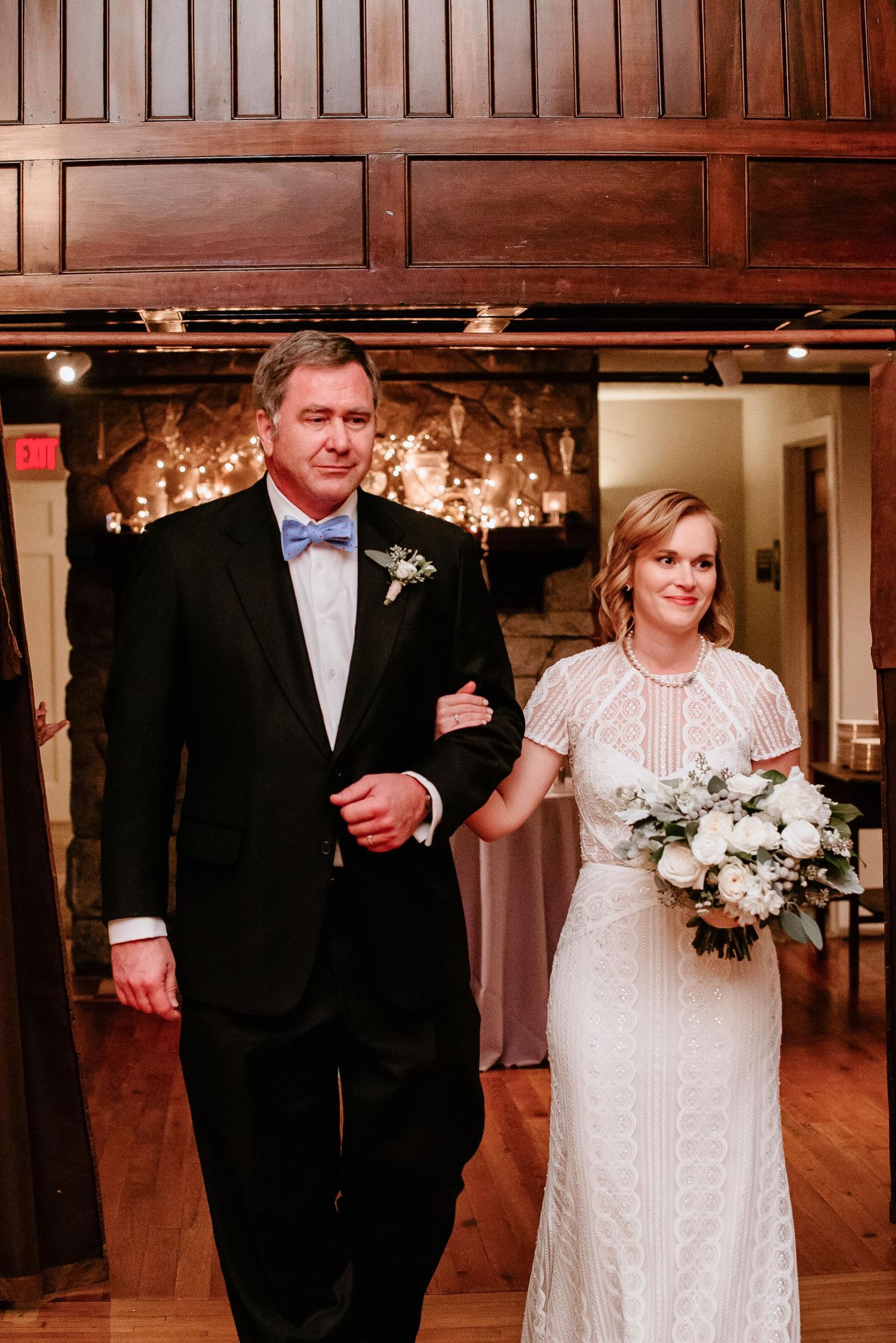 Bride in lace gown holding white bouquet walks down aisle with man in black tuxedo and blue bow tie.