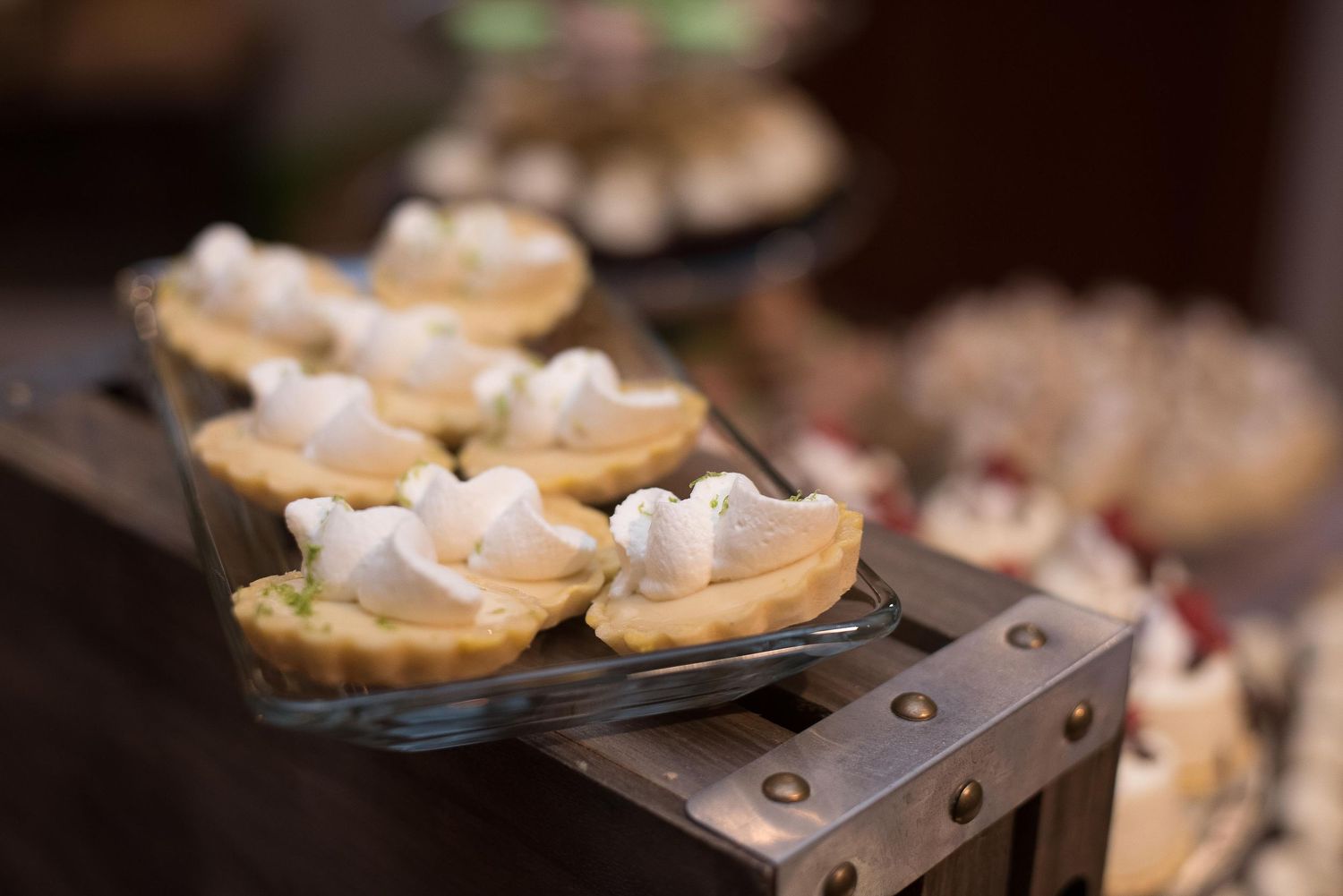 Mini lemon meringue tarts with whipped cream topping displayed on a glass tray at a dessert table.