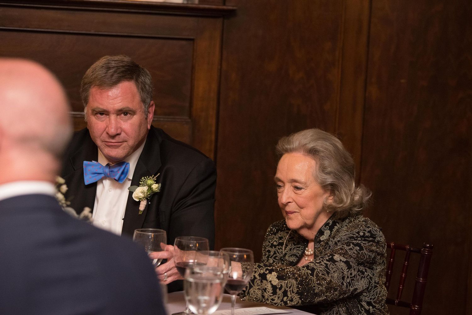 Elegantly dressed man in bow tie and woman in black seated at formal dinner table with glassware.