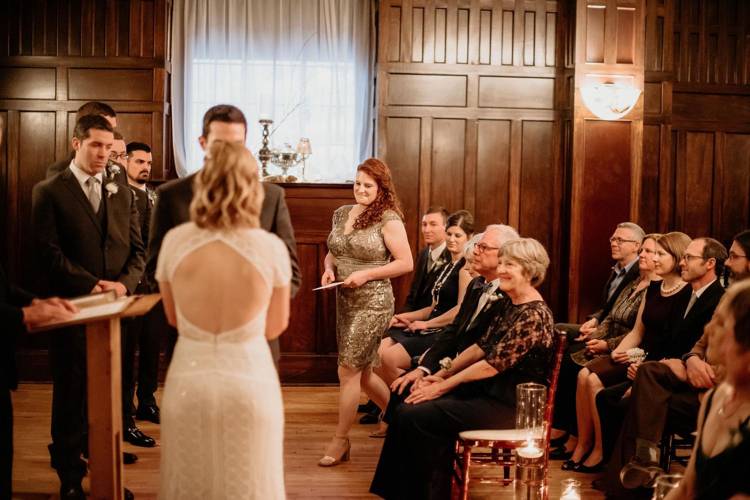 Wedding ceremony in wood-paneled room with bride, groom, and officiant at altar while seated guests watch attentively.
