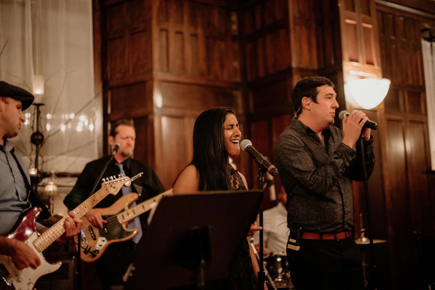 Live band performing on stage with guitarist and two vocalists singing into microphones in elegant wood-paneled venue.