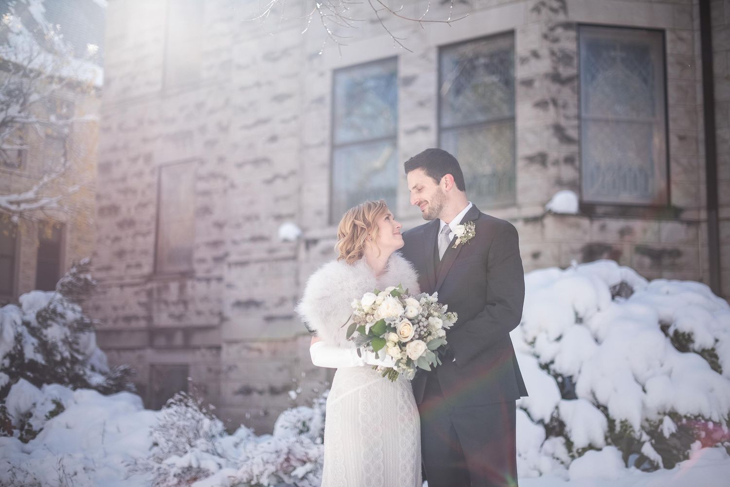 A couple shares a romantic winter wedding moment outdoors, surrounded by snow-covered bushes near a stone building.