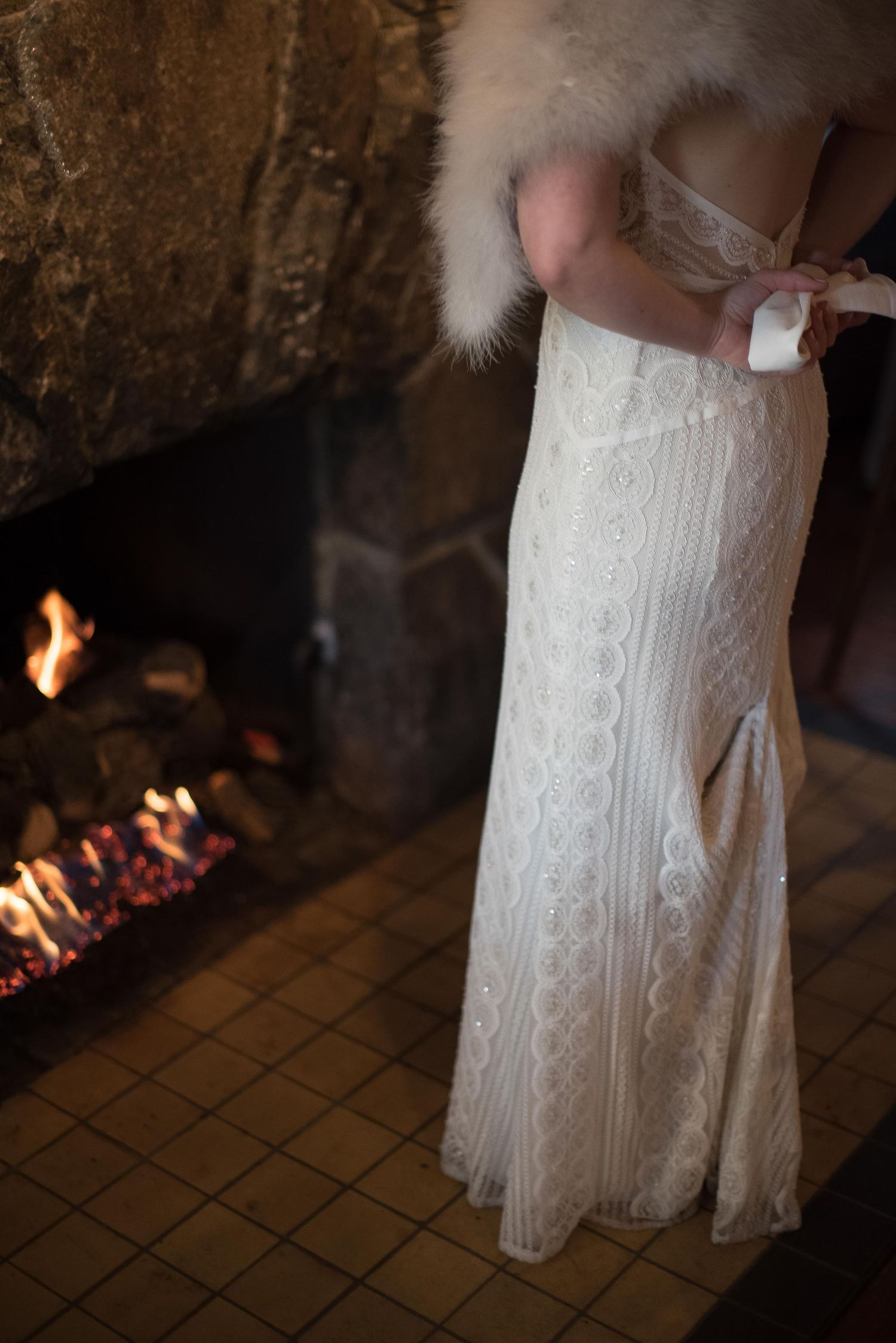 Bride in fitted lace wedding gown with fur stole being buttoned near a stone fireplace.