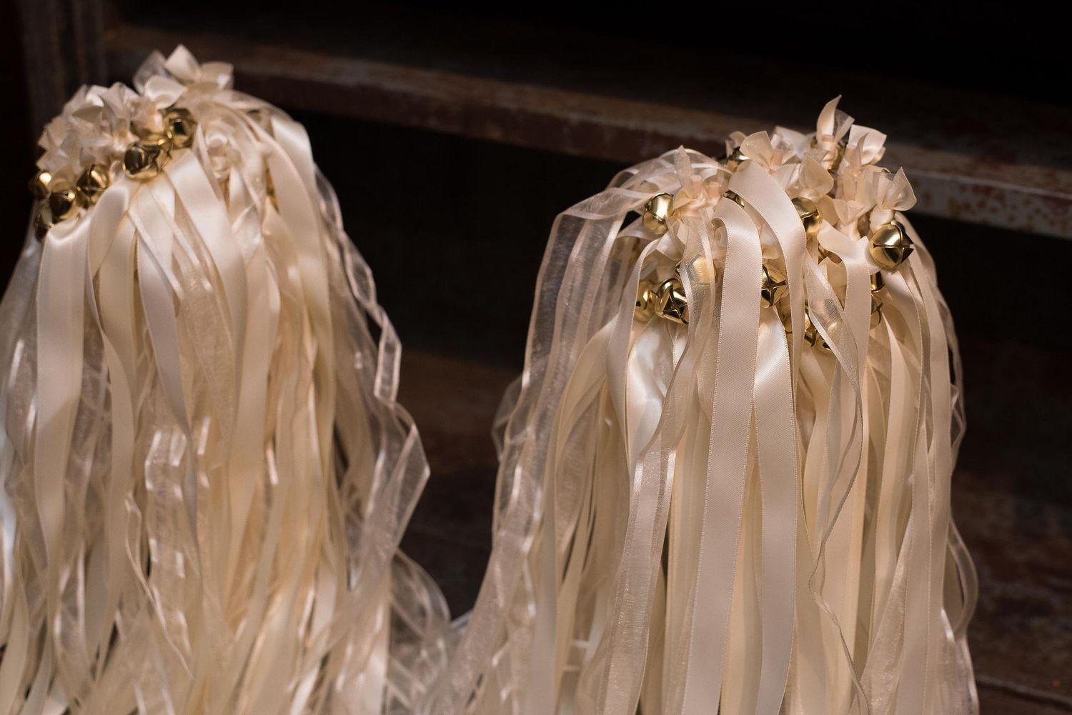 Two elegant wedding wands with cream satin ribbons and gold bell accents displayed against a wooden backdrop.