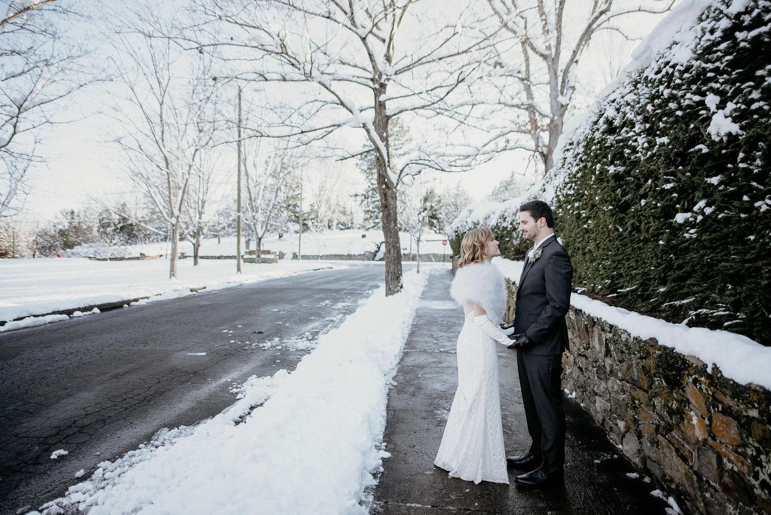 Bride and groom share a romantic moment beside a snowy stone wall on their winter wedding day.