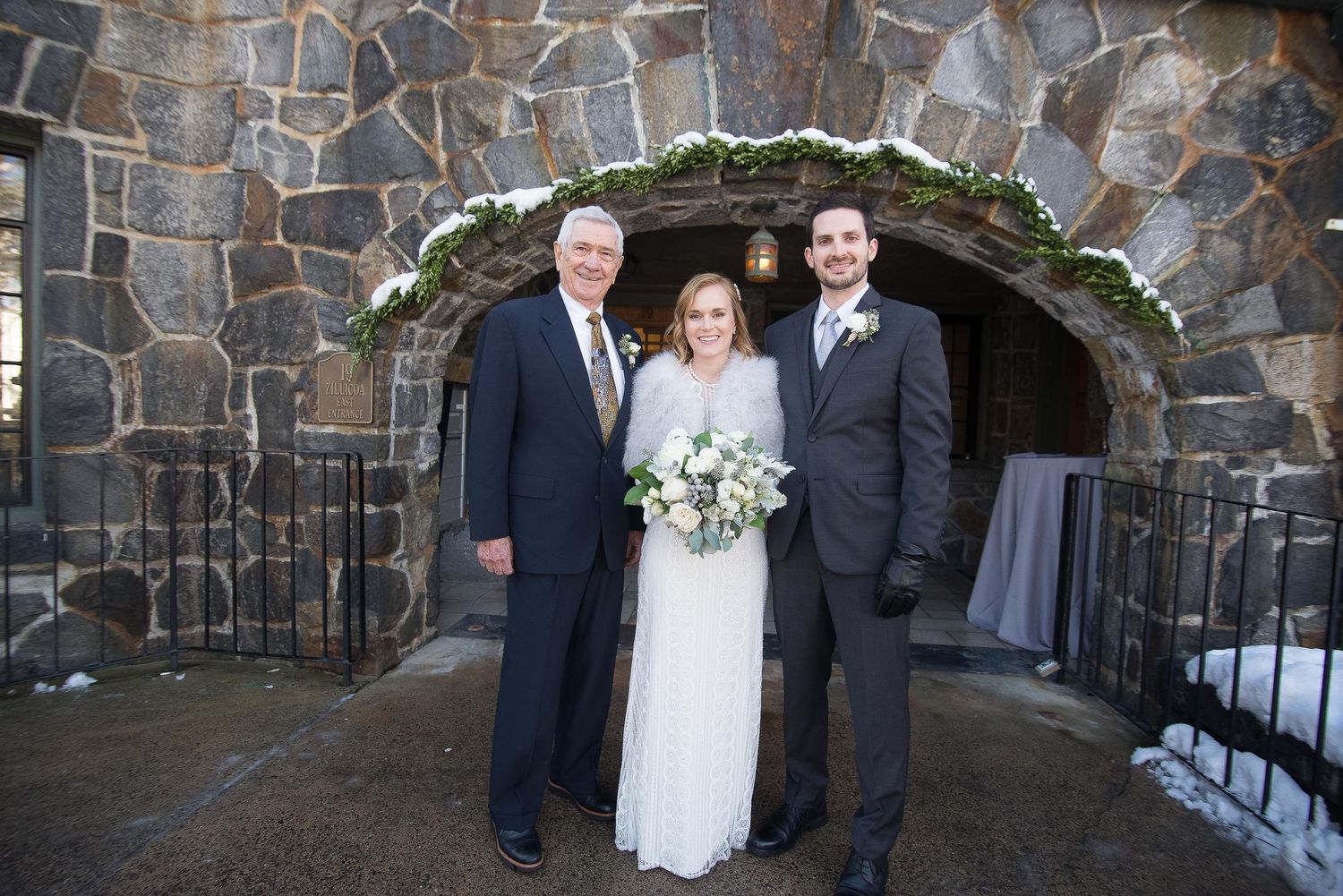 Wedding couple and male guest smile together under snow-dusted stone archway at elegant winter wedding ceremony.