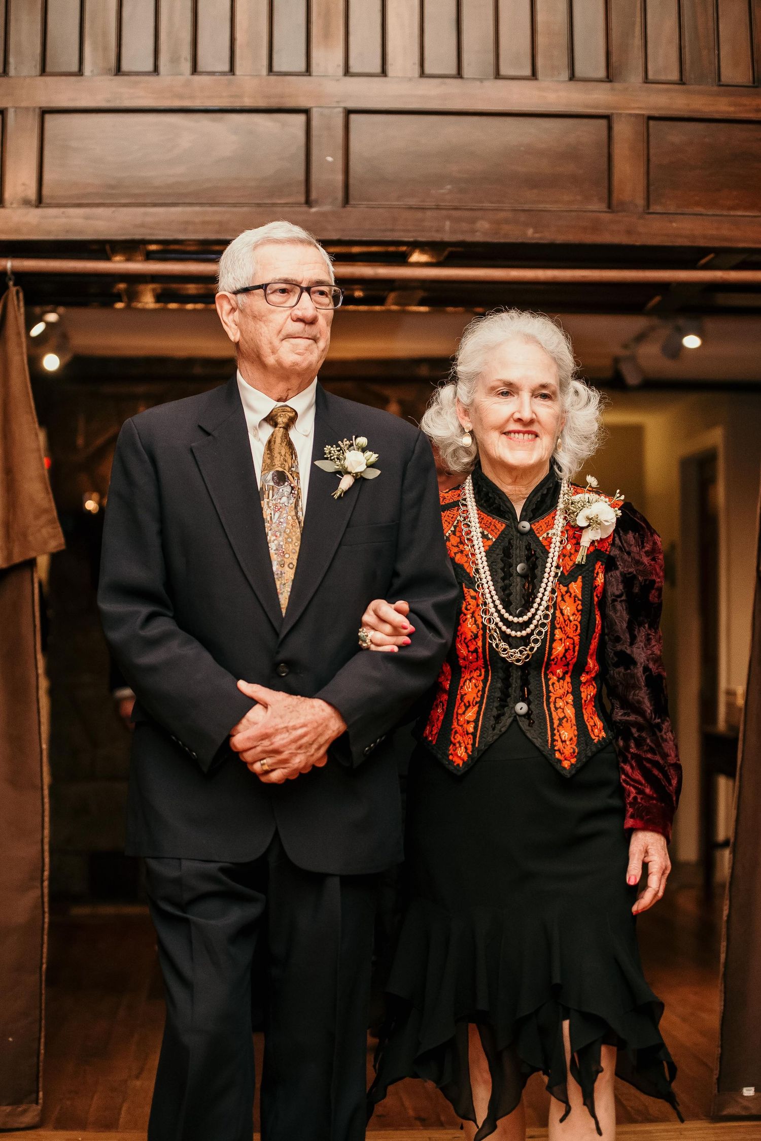 Elderly couple dressed formally in black attire with boutonnieres, posing together at an elegant indoor event.
