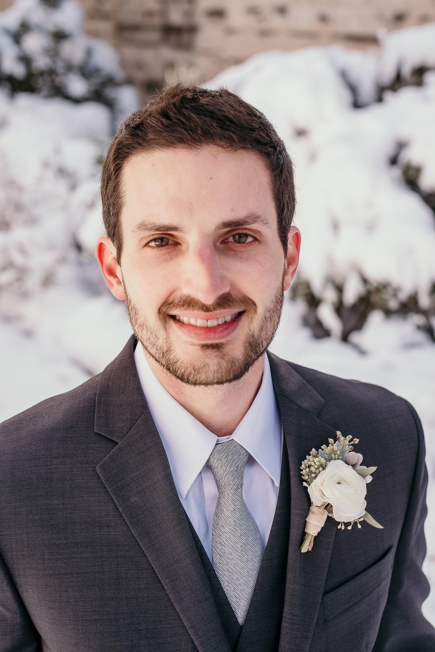 Smiling groom in gray suit with white boutonniere posing outdoors in snowy winter setting.