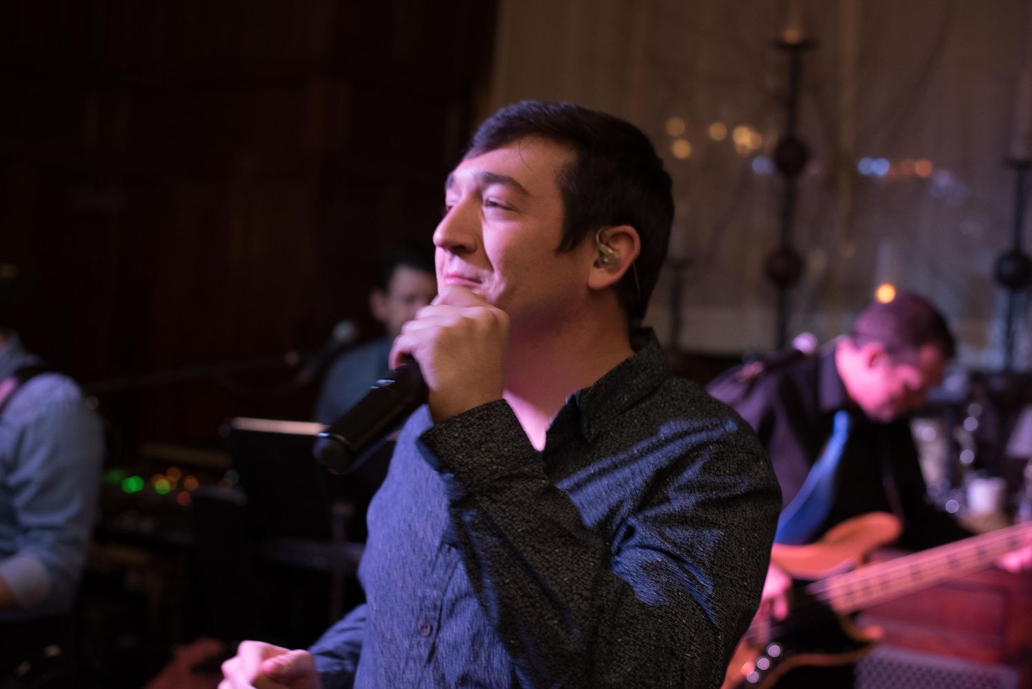 A young man smiles with eyes closed at a live music event, with band members visible in the background.