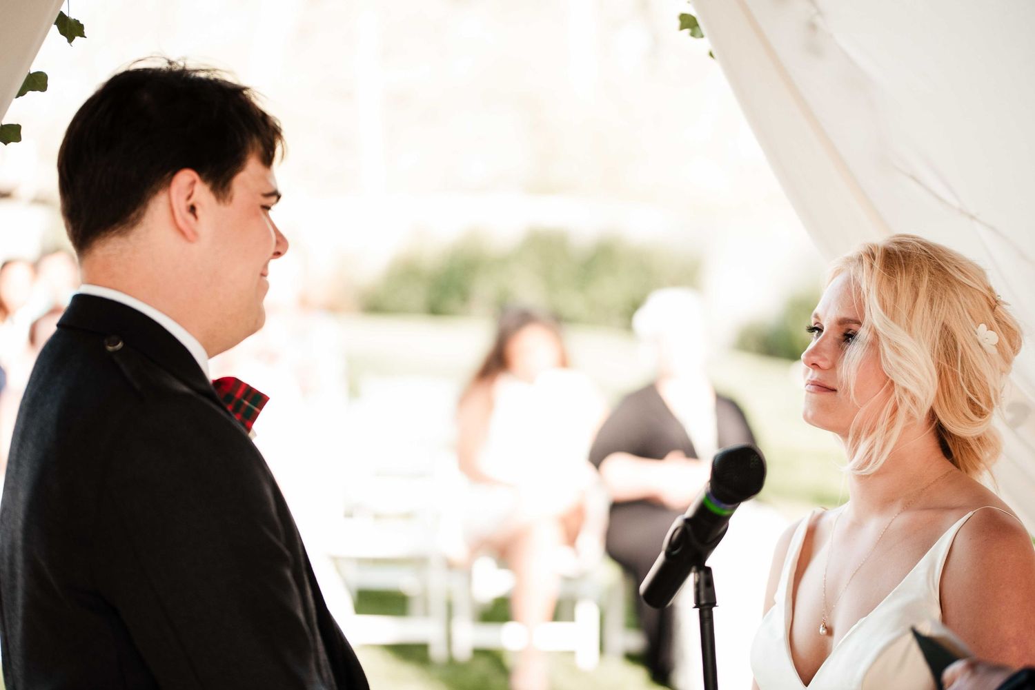 Bride and groom exchange wedding vows at an outdoor ceremony under a white tent.