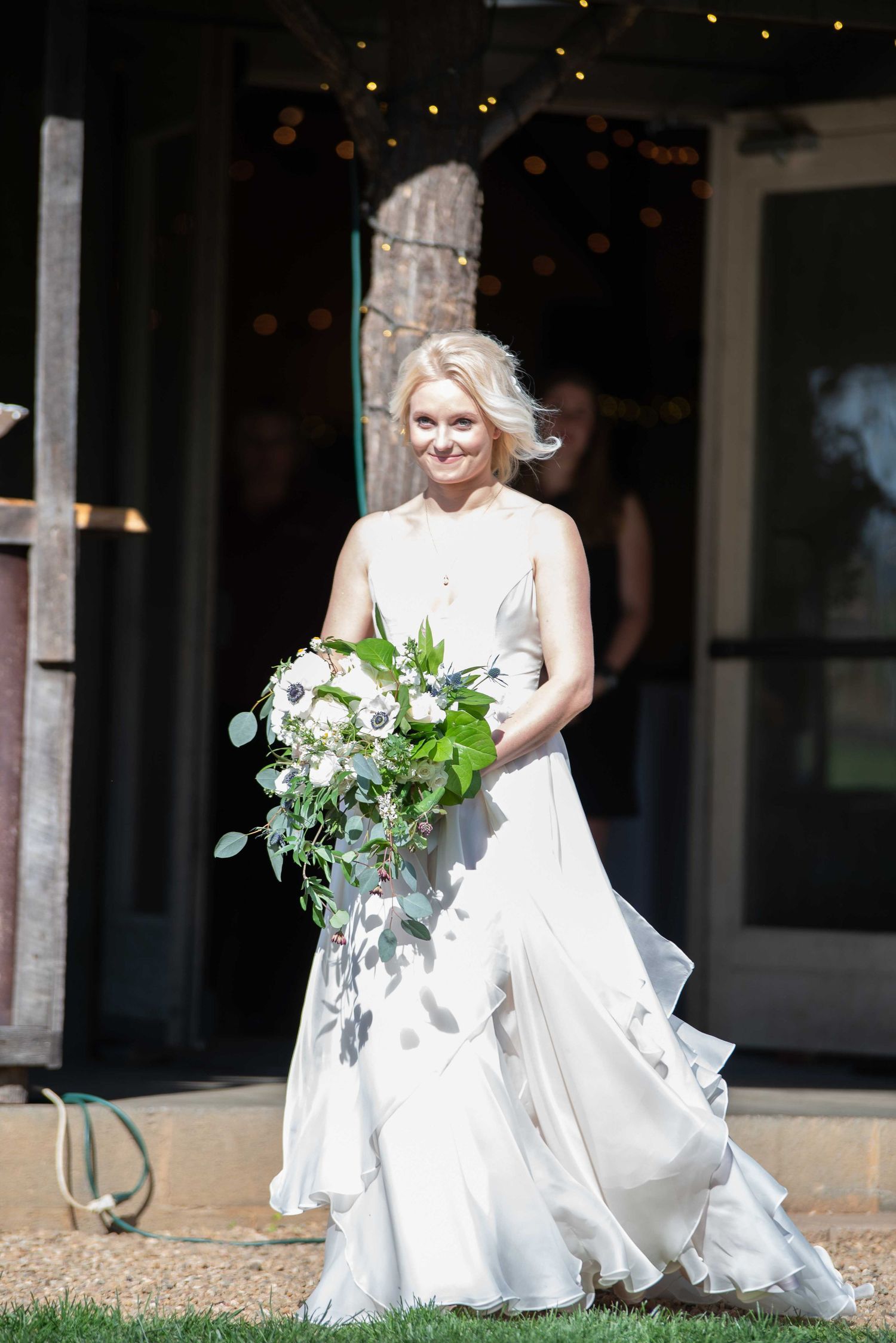 Smiling bride in white ruffled gown holding large greenery bouquet walking outdoors on sunny wedding day.