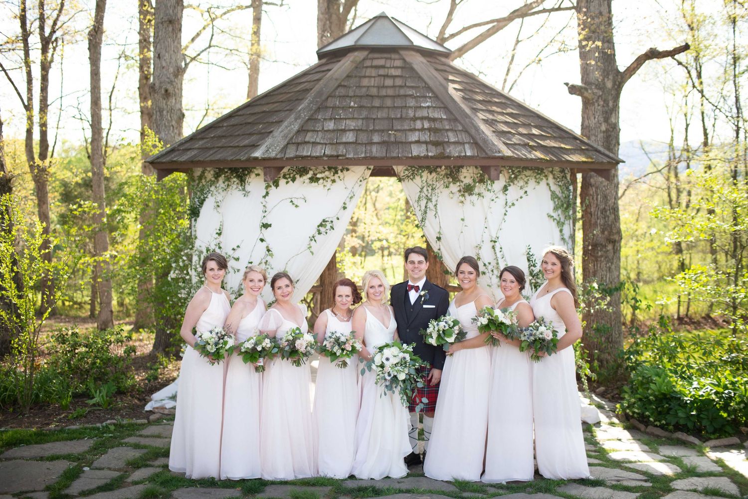 Wedding party poses under floral gazebo, bridesmaids in blush gowns with bouquets, groom in kilt, lush green garden backdrop.