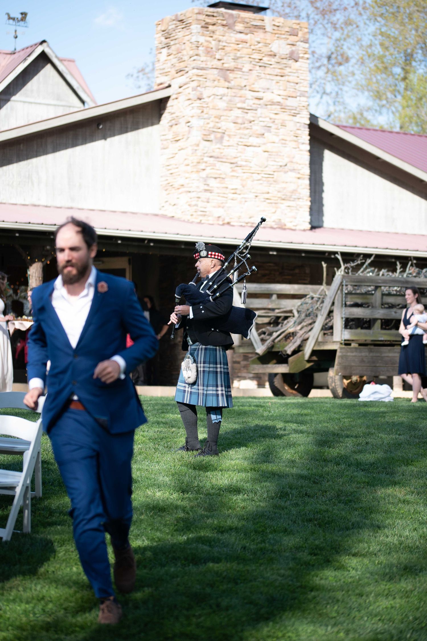 A bagpiper in traditional Scottish attire performs outdoors at a rustic barn wedding ceremony on a sunny day.