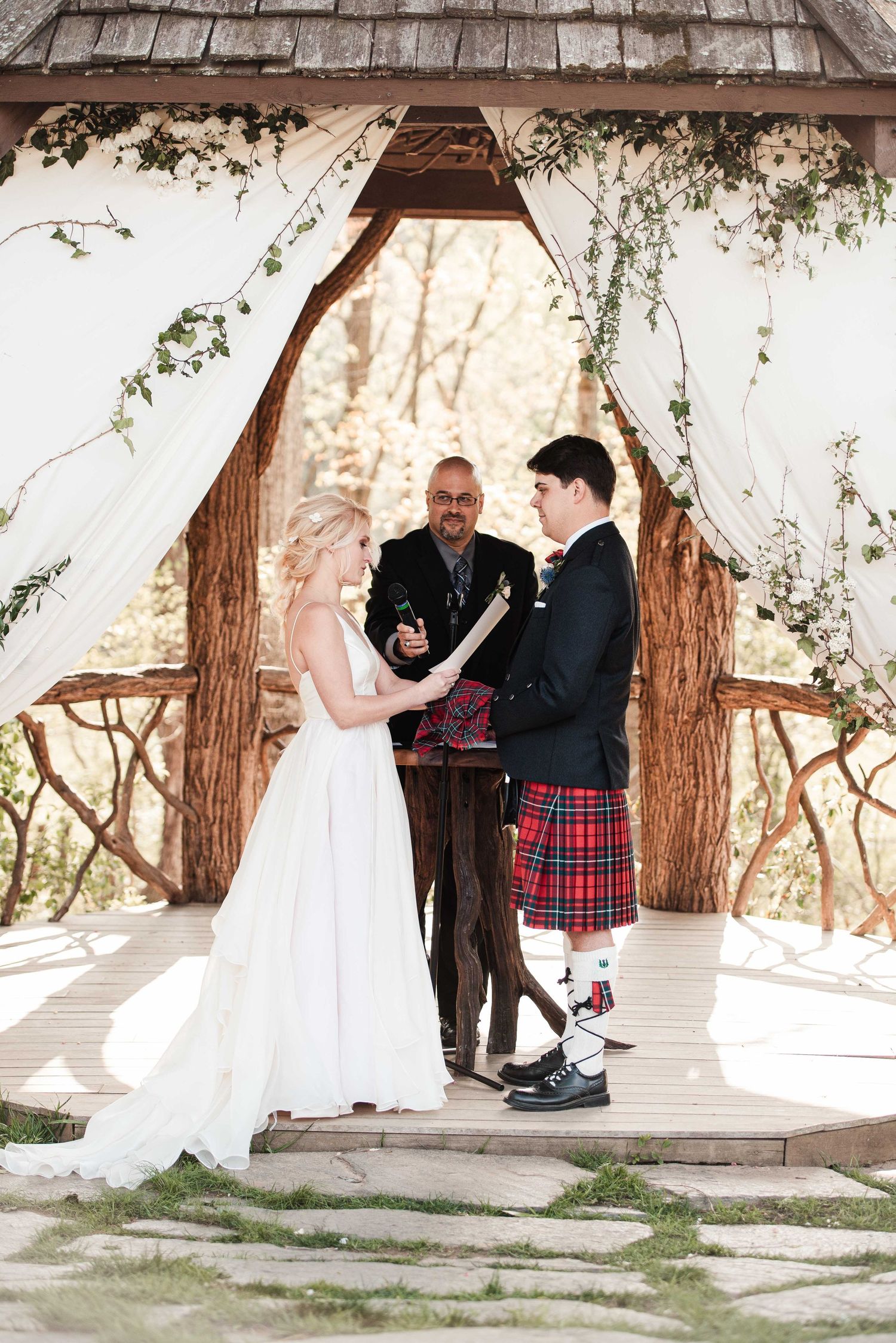 Bride and groom exchange vows under rustic wooden arch with white draping, officiant presiding at outdoor wedding ceremony.