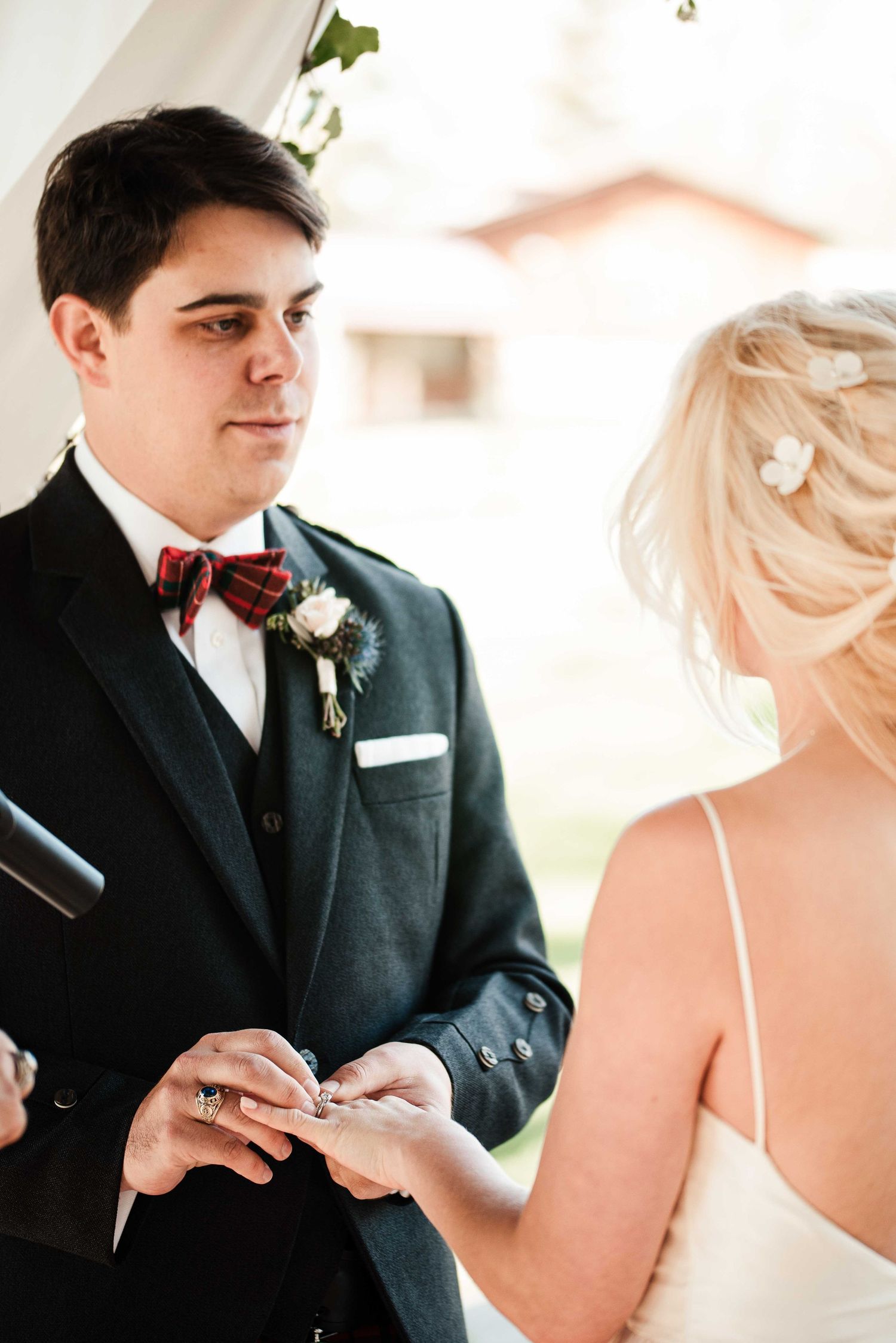 Groom in dark suit with plaid bow tie places wedding ring on bride's finger during outdoor ceremony.