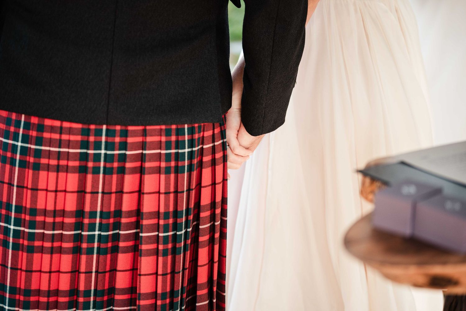 Close-up of a red tartan kilt and white wedding dress, with couple holding hands showing a ring.