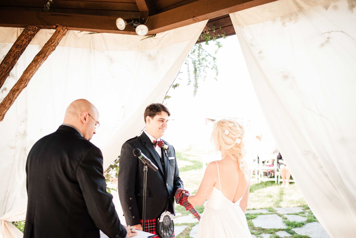 Bride and groom exchanging vows at a rustic outdoor wedding altar with white draping and officiant.