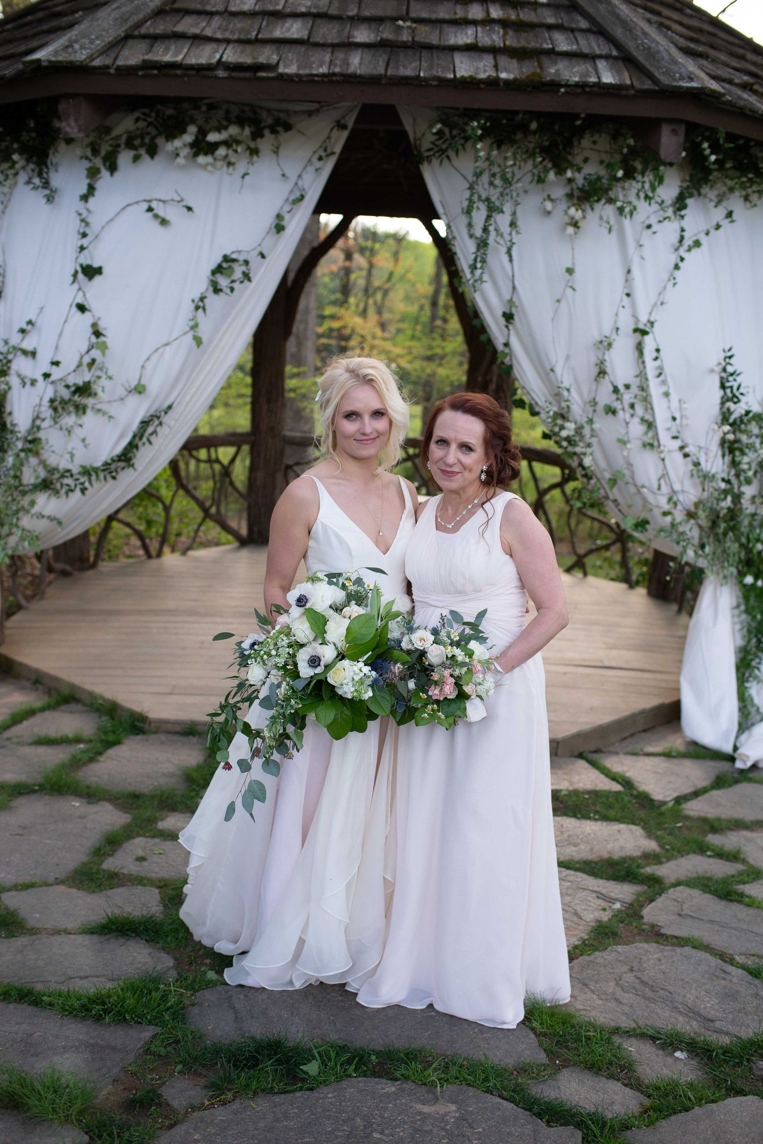 Two women in white gowns holding bouquets pose under a floral gazebo at an outdoor wedding ceremony.