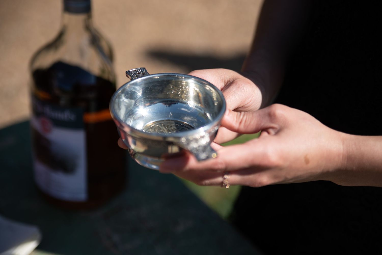 Hands holding a silver quaich cup with a bottle of Scotch whisky in the background at a Scottish wedding.