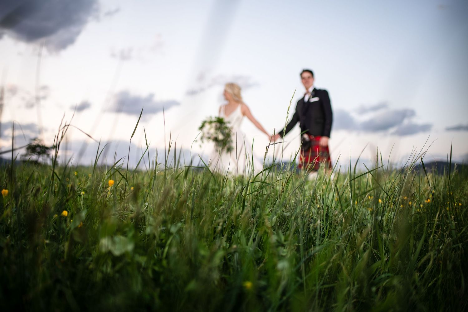 Bride and groom walk hand in hand through a lush green meadow under a dramatic cloudy sky.