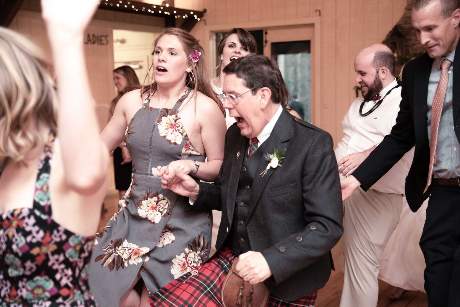 Guests dancing energetically at a wedding reception, wearing formal attire including a Scottish kilt.