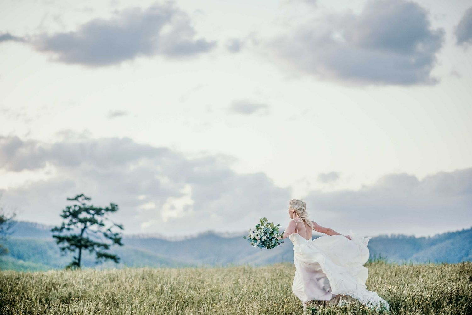 Bride in flowing white gown holding bouquet runs through green meadow with mountain backdrop and cloudy sky.