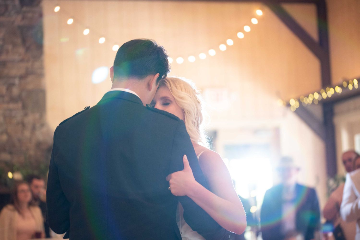 Bride and groom share first dance embrace in rustic barn venue with warm string lights glowing overhead.