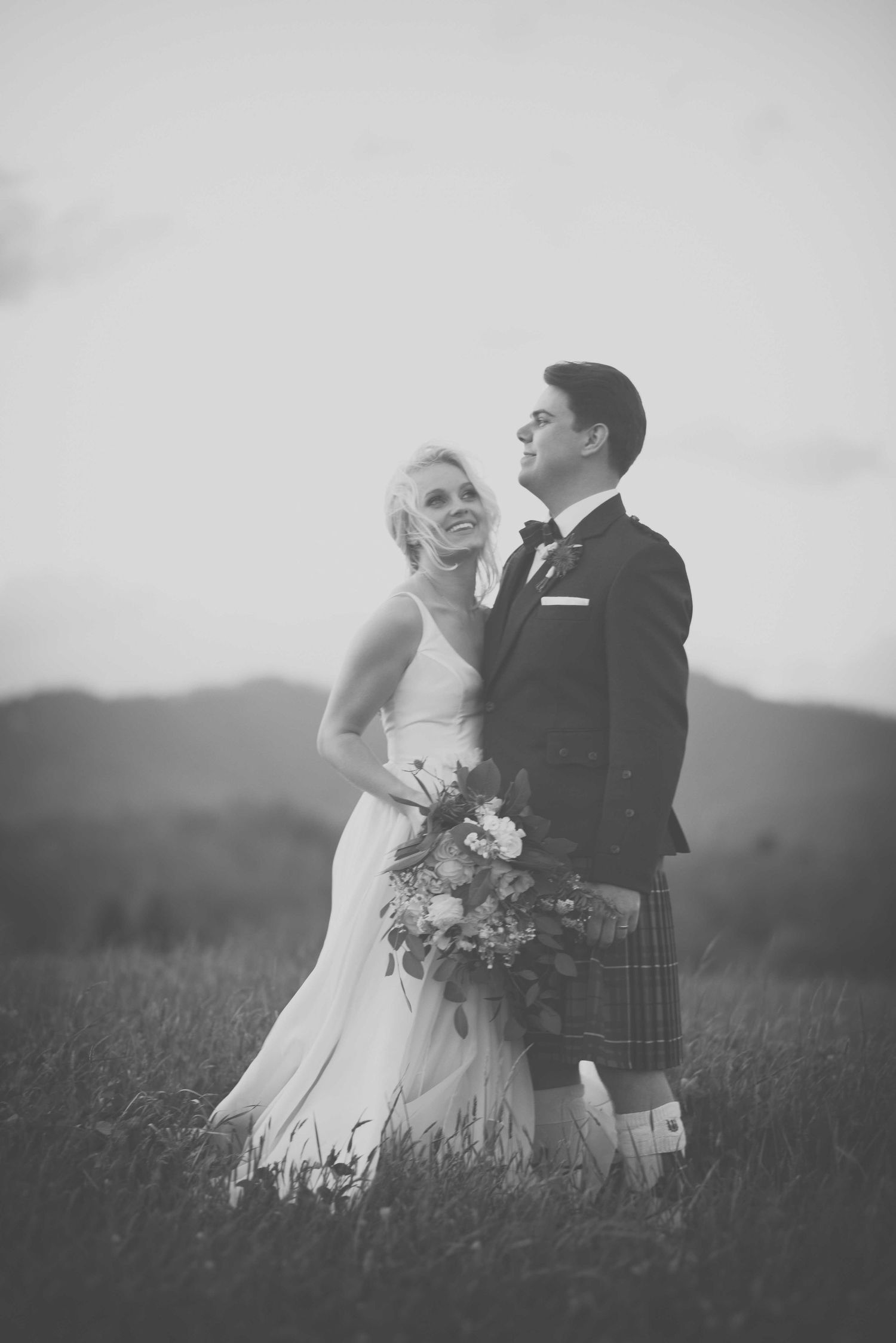 Black and white photo of a bride and groom in a field, groom wearing a kilt, bride holding a bouquet.