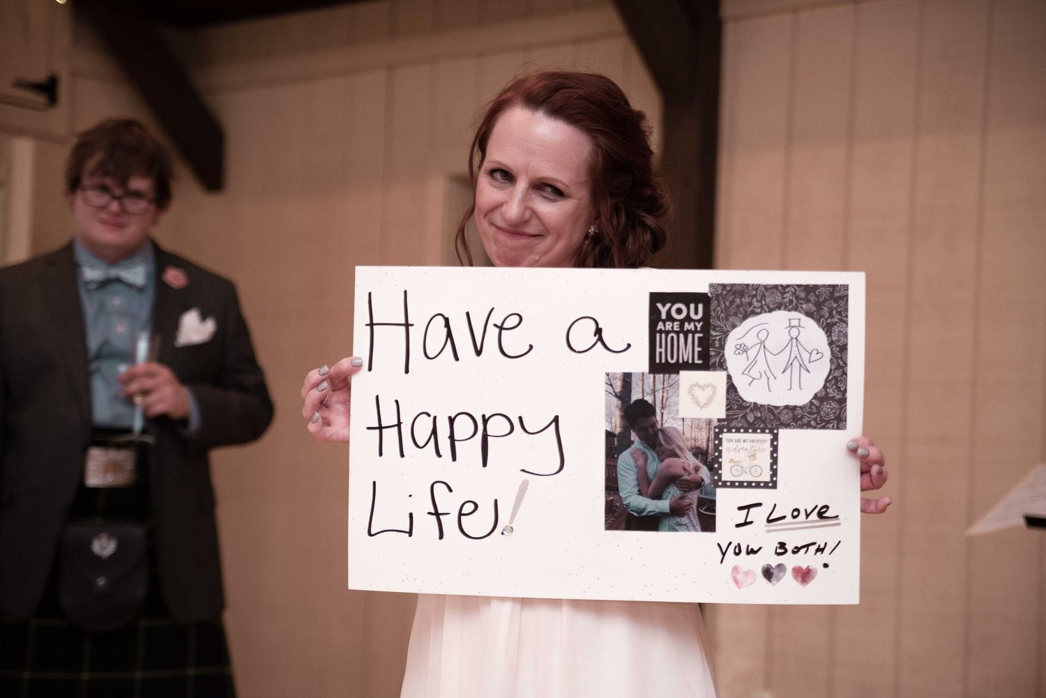 Bride holding handmade sign reading Have a Happy Life I Love You Both at wedding reception.